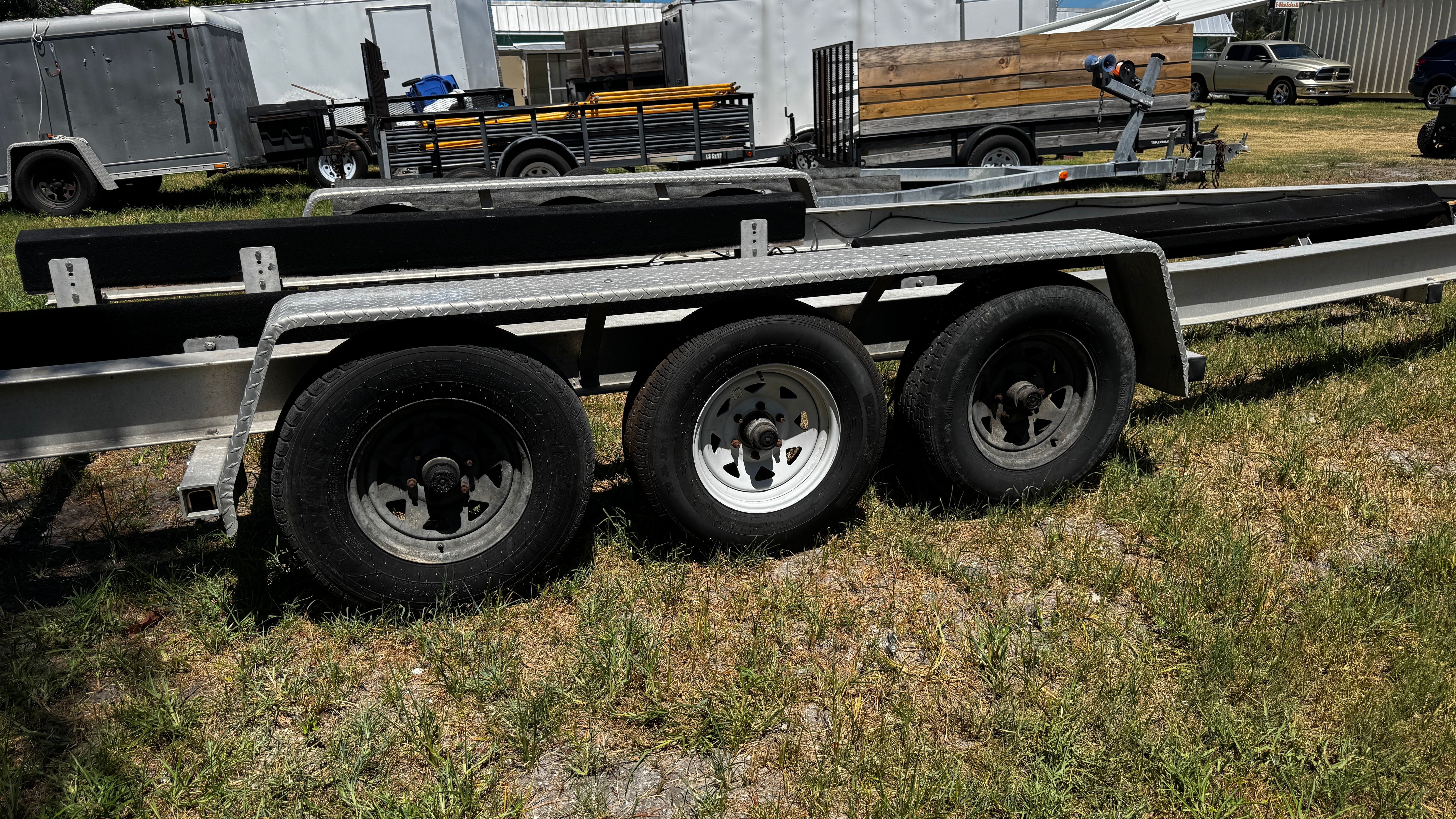 A triple-axle boat trailer on grass, surrounded by trailers and vehicles.