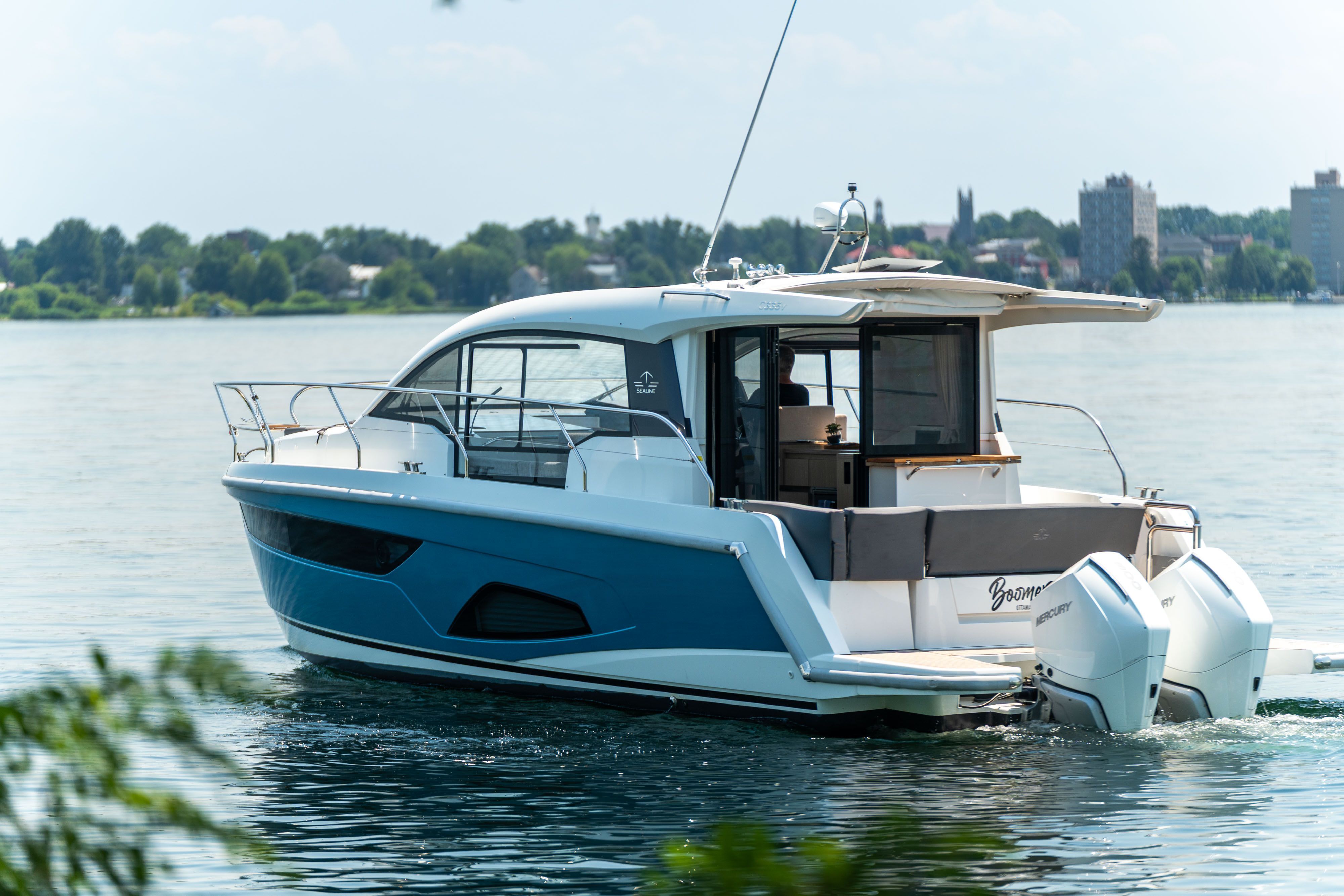 A modern Sealine C335V yacht cruising on calm water with a cityscape in the background.