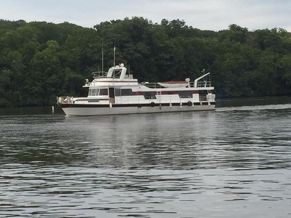 1980 Pluckebaum Baymaster yacht cruising on a calm river with lush green forest backdrop.