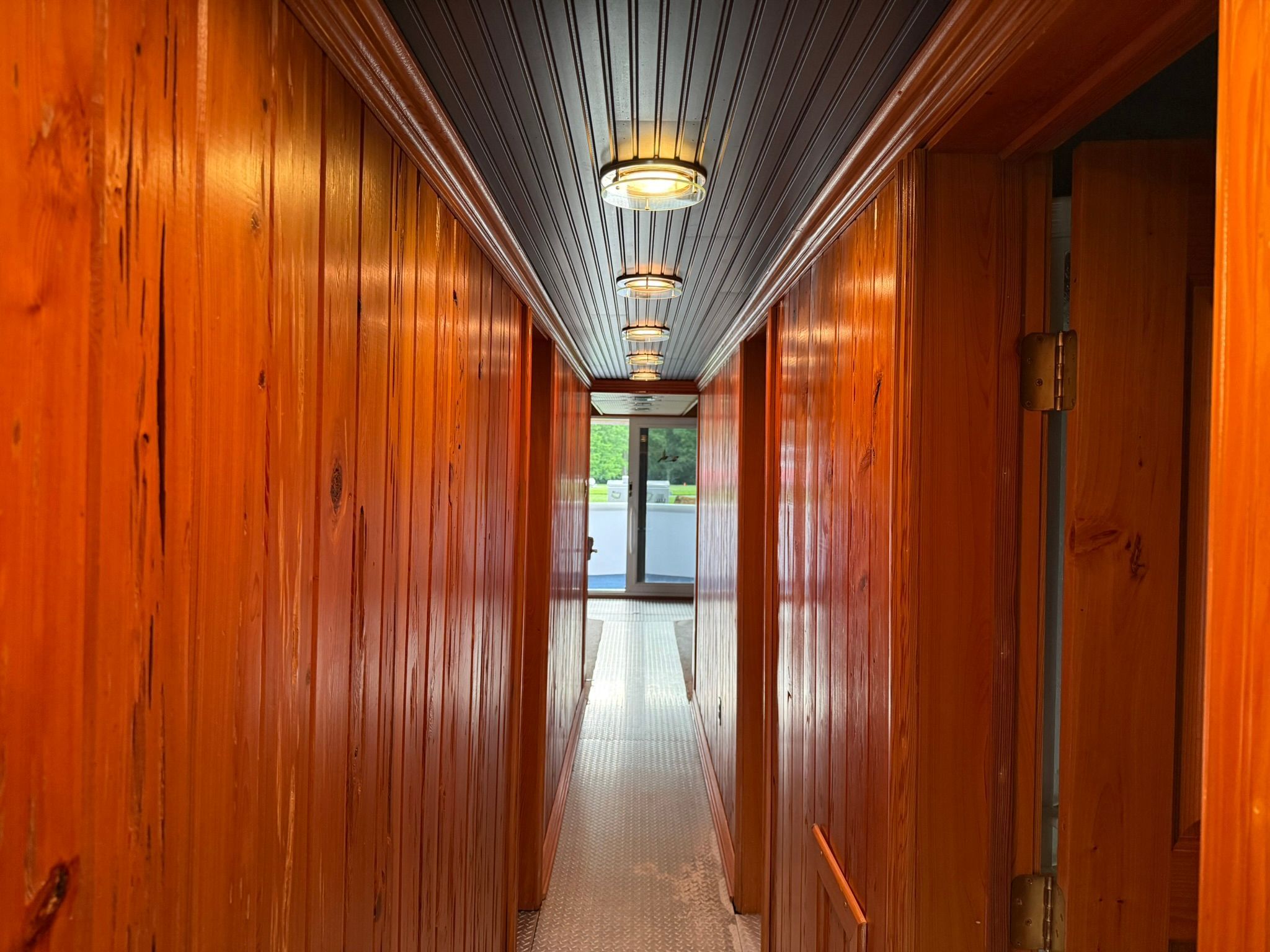 A narrow, wood-paneled hallway with round ceiling lights leads to a window overlooking the outside.