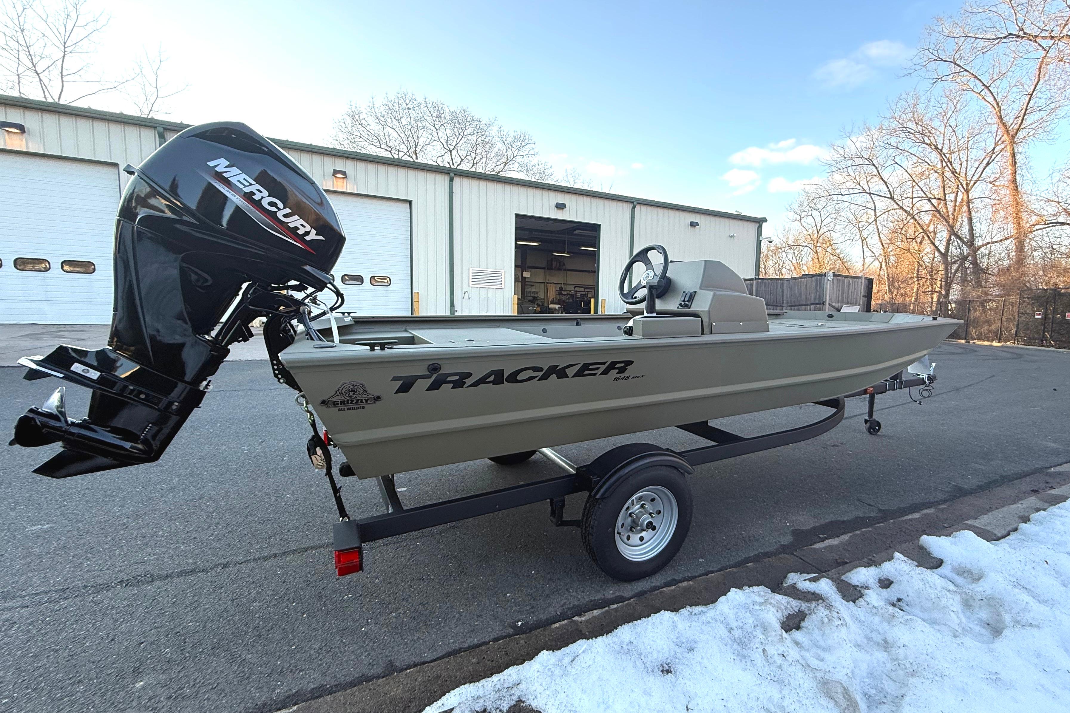 2026 Tracker Grizzly 1648 SC boat with Mercury engine on trailer, parked outside a garage.