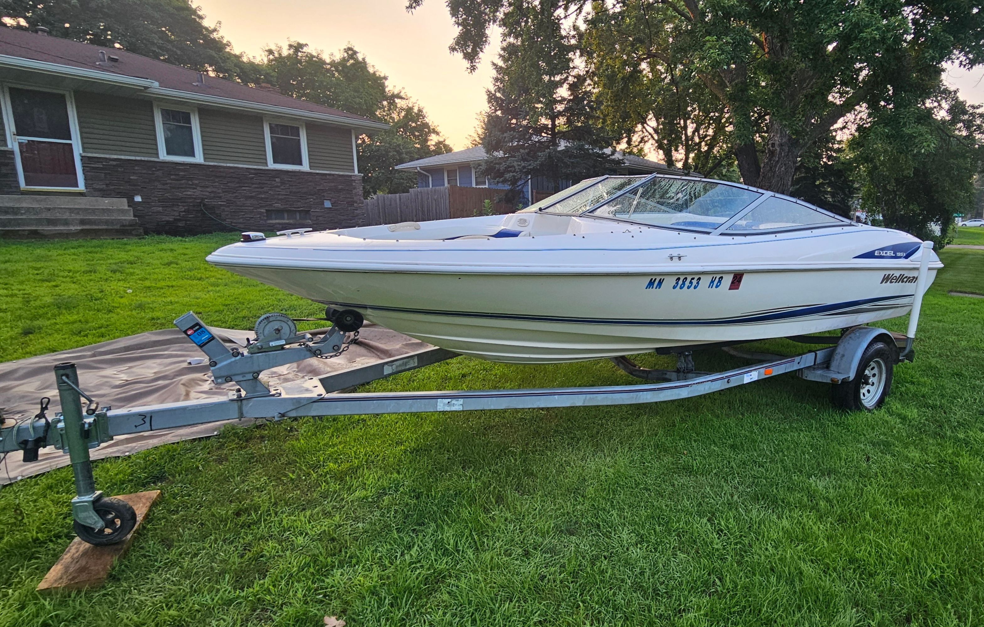 A 1996 Wellcraft Excel boat on a trailer sits on a grassy lawn in front of a house.