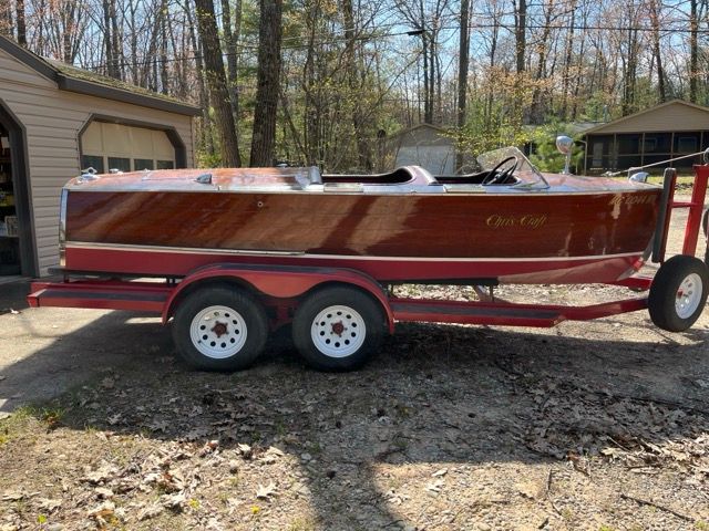 A 1938 Chris-Craft Custom Deluxe Runabout on a trailer in a wooded area.