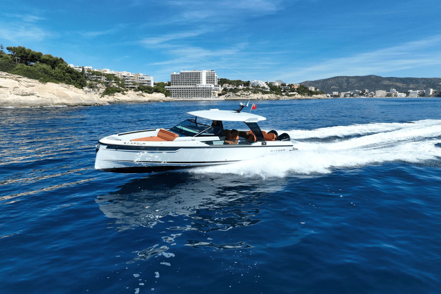 A Saxdor SX320 GTO boat speeds across the sea, with coastline and buildings in the background.