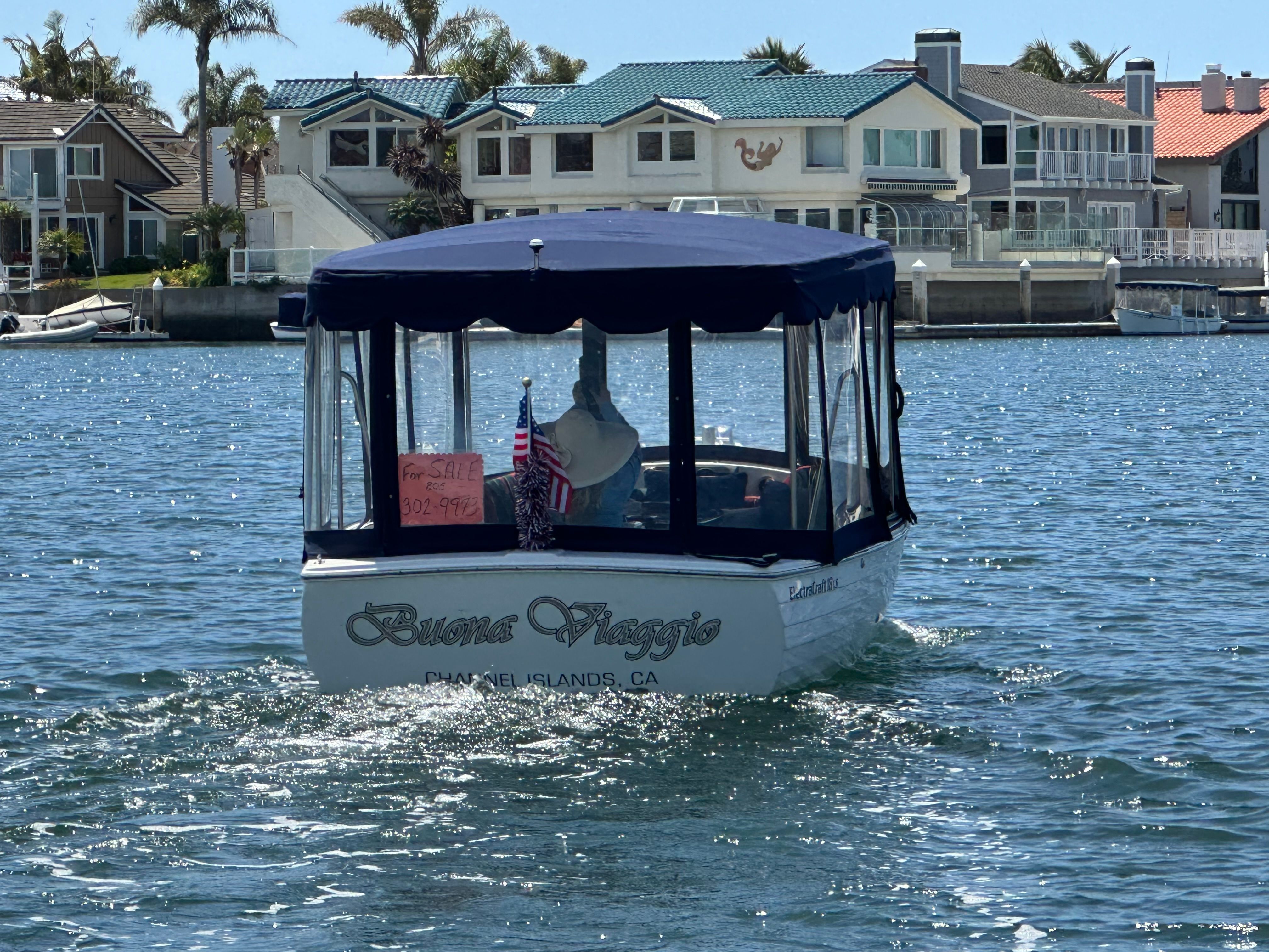 A white boat with a blue canopy cruises on calm water in front of elegant houses.
