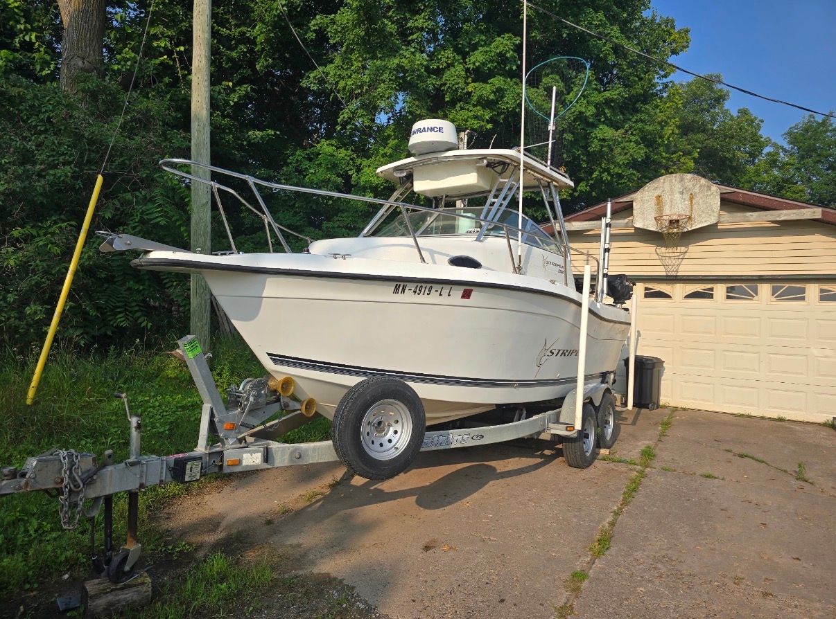 A white boat on a trailer in front of a garage, surrounded by trees.