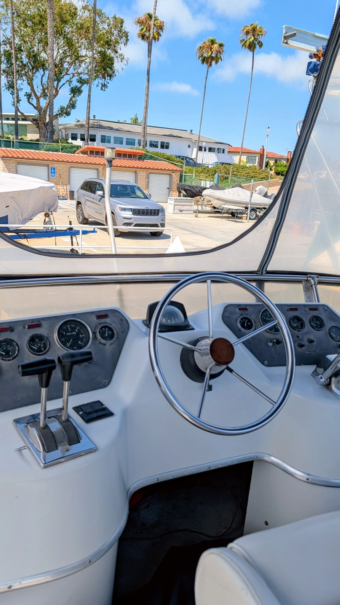 The image shows the helm of a 1987 Chris-Craft 320 Amerosport Express. There is a steering wheel, gauges, and levers. Boats and a car are visible in the background.