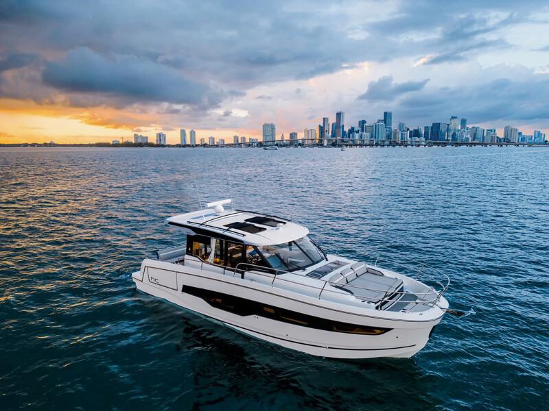 2025 Jeanneau NC 1295 Coupe yacht cruising near a city skyline at sunset.