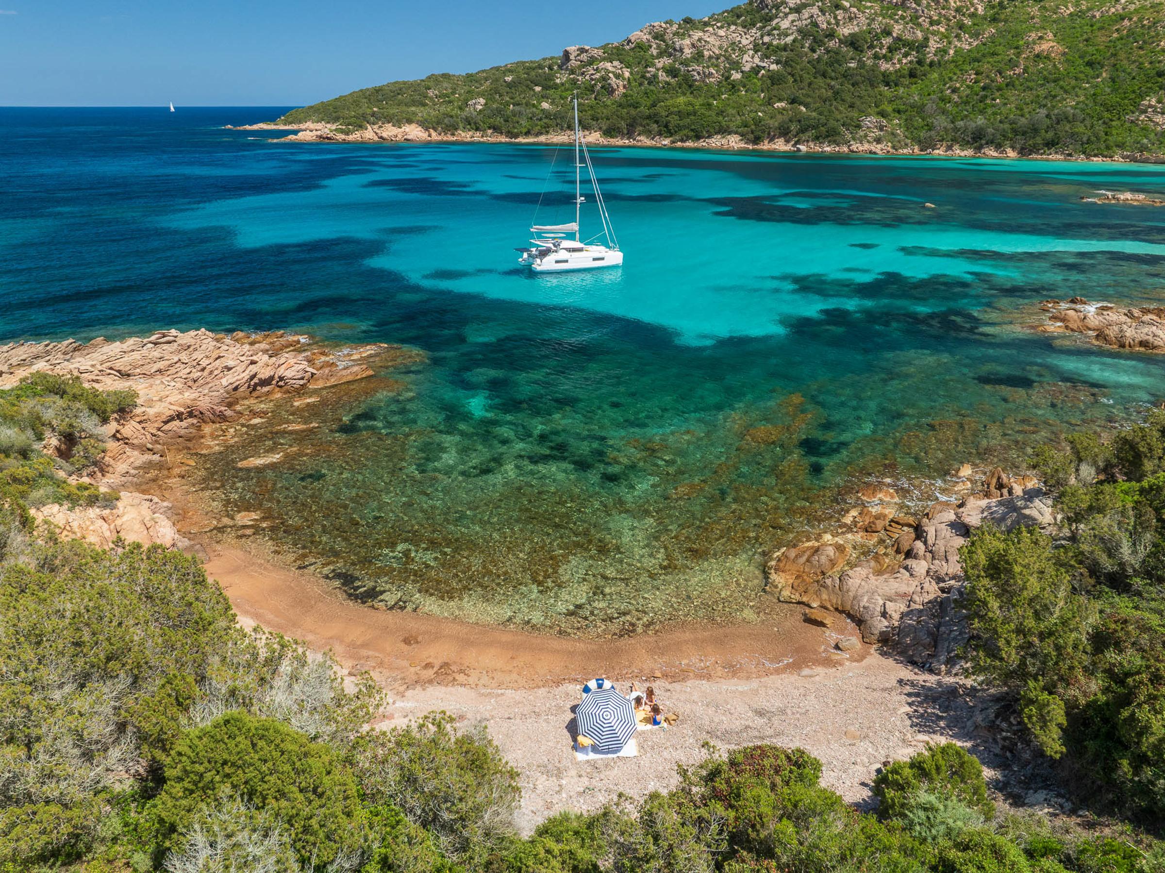 A Lagoon 38 catamaran anchored in a serene turquoise bay, surrounded by lush greenery and rocky shores.