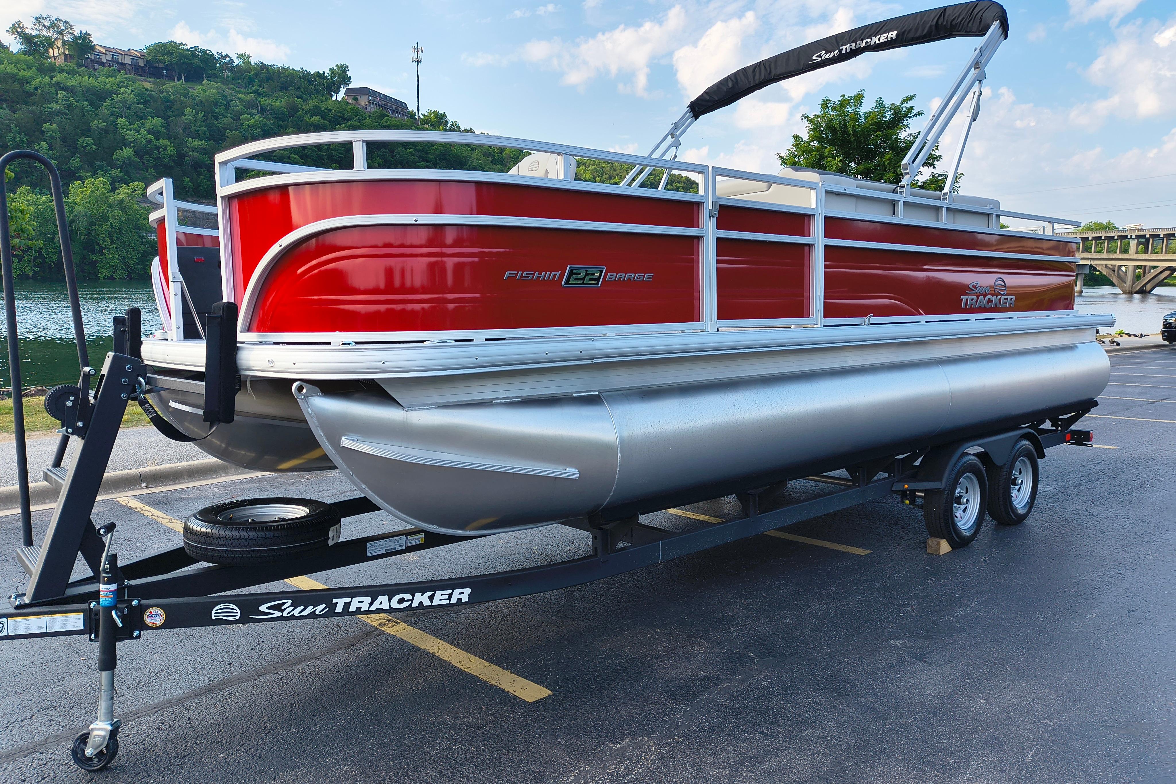 2026 Sun Tracker Fishin' Barge 22 DLX pontoon boat on trailer by a river.