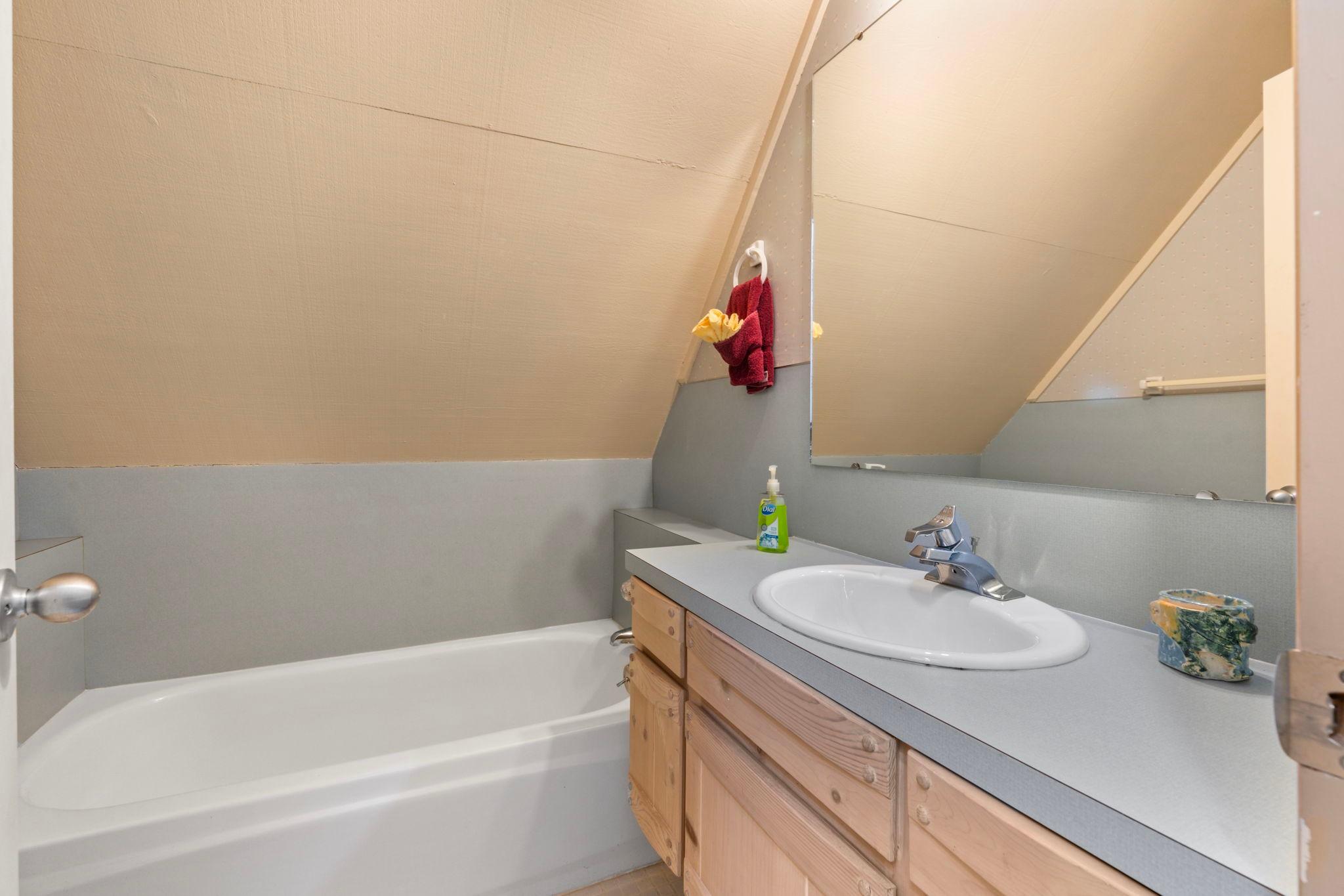 Small bathroom with angled ceiling, bathtub, wooden vanity, and large mirror in a 1976 custom boathouse.