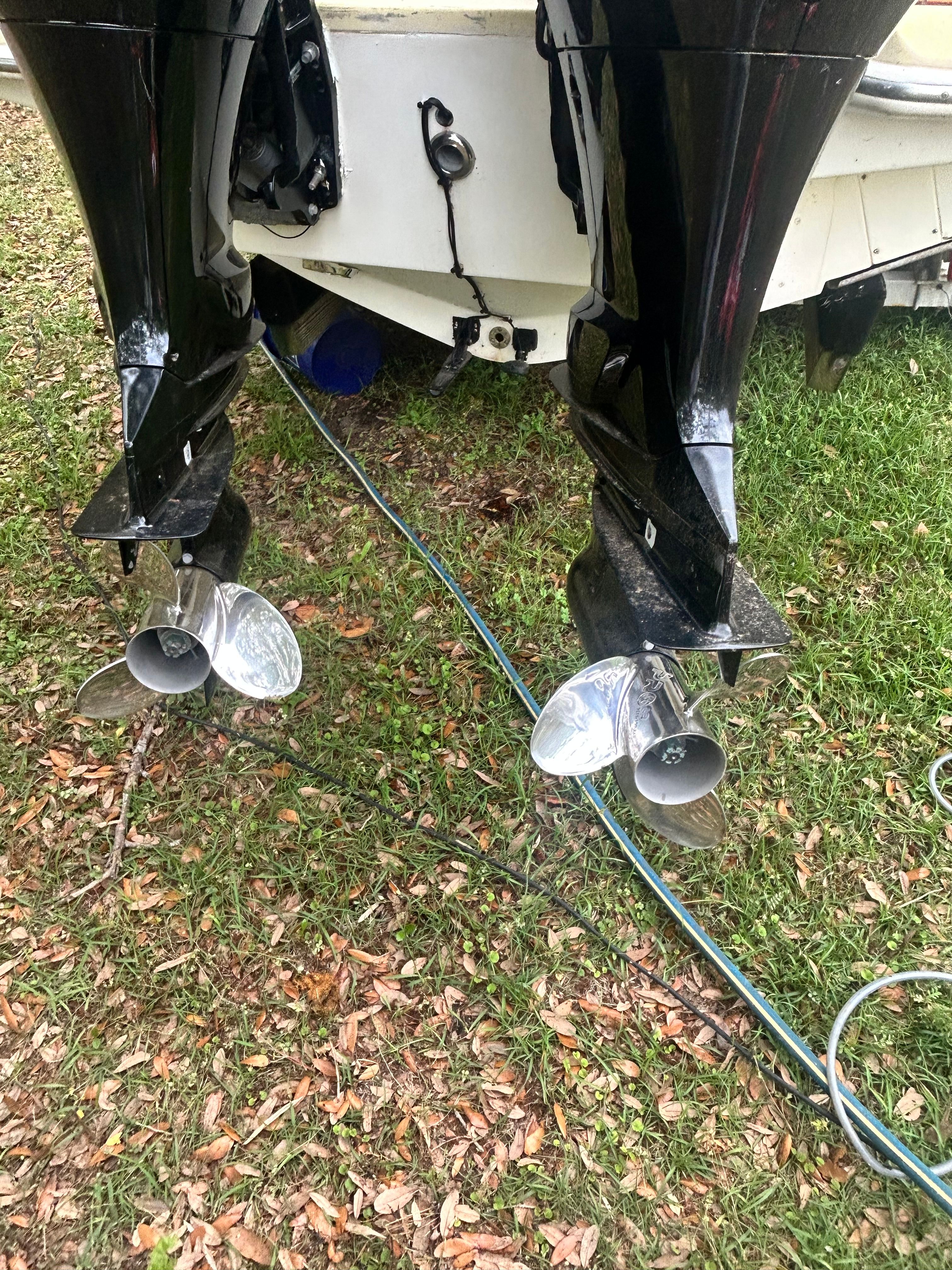 Two outboard motors with silver propellers are attached to a boat resting on grass.