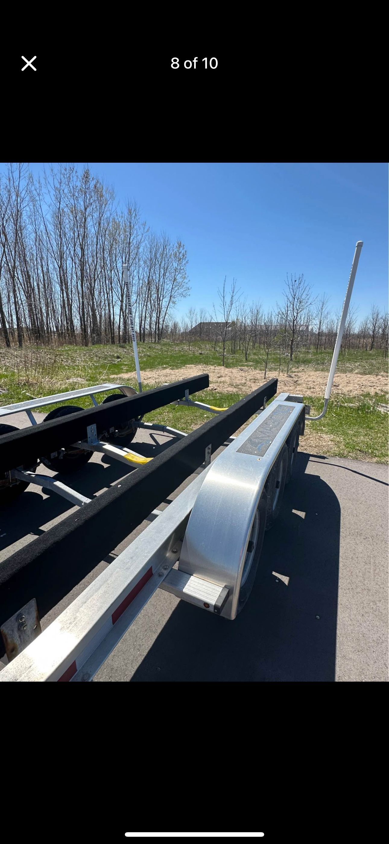A boat trailer is parked on a lot, surrounded by bare trees and a clear blue sky.