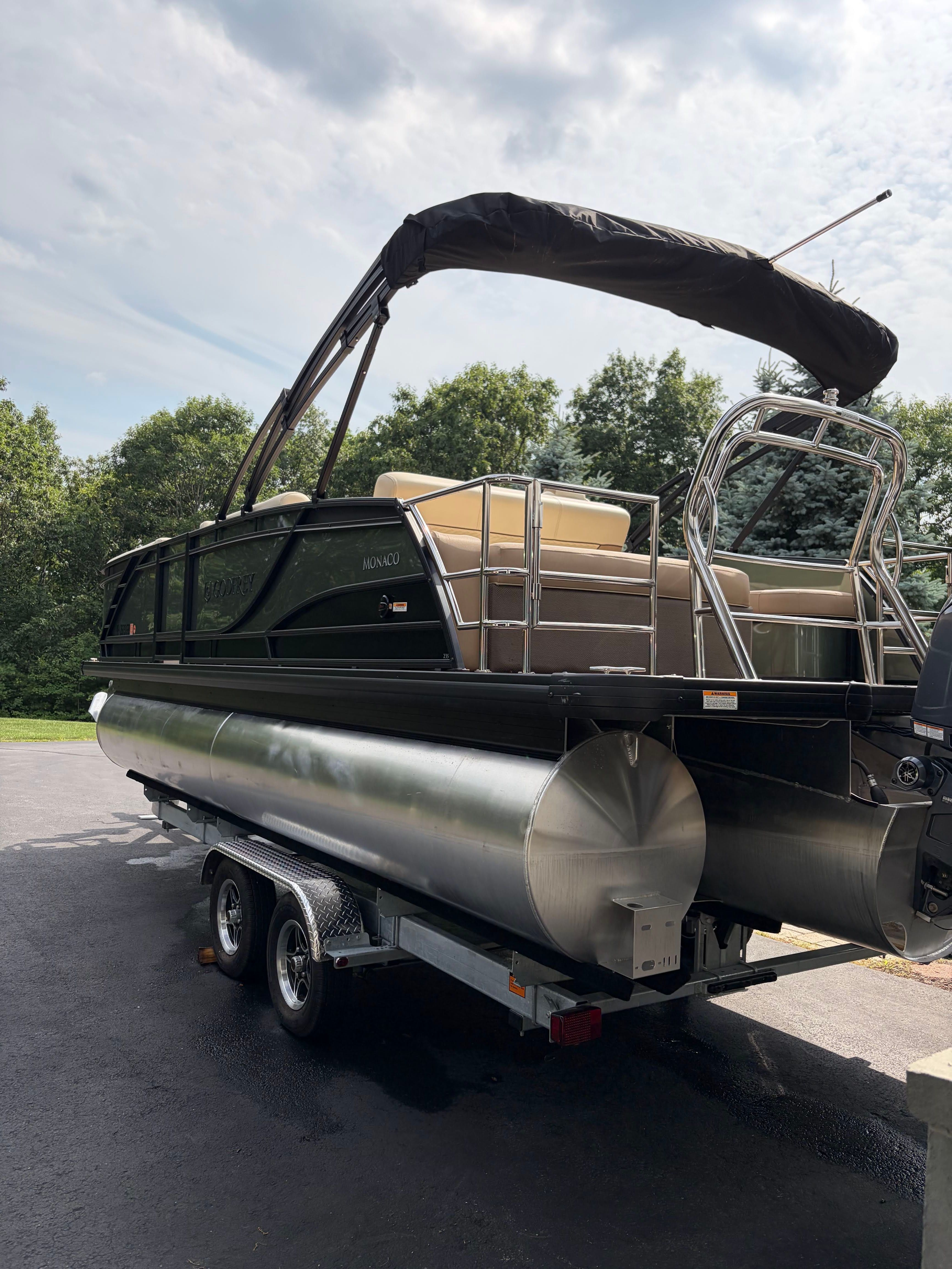 A 2025 Godfrey Monaco Cruise pontoon boat on a trailer, surrounded by trees.
