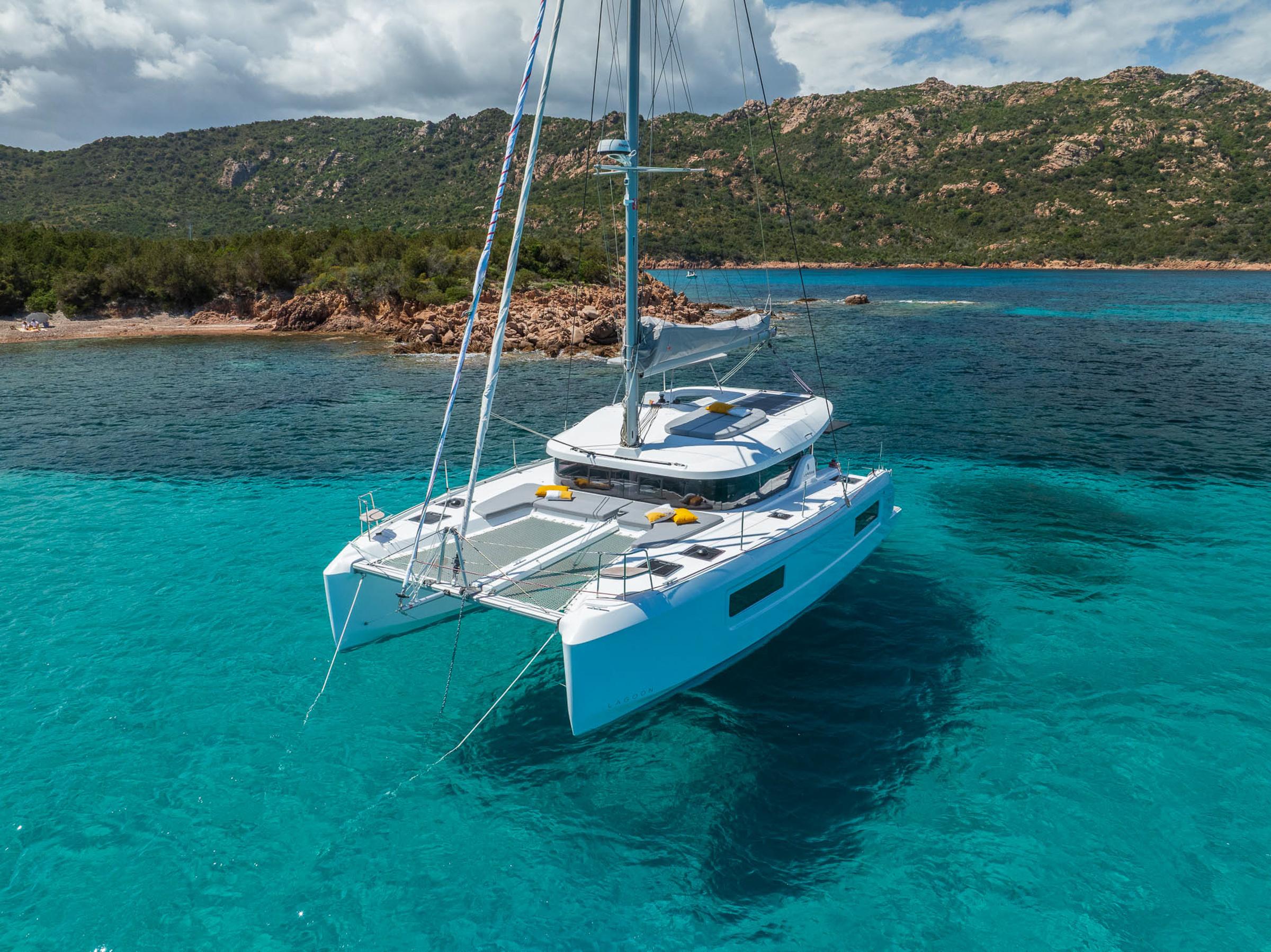 2025 Lagoon 38 catamaran anchored in clear turquoise waters near a rocky coastline.