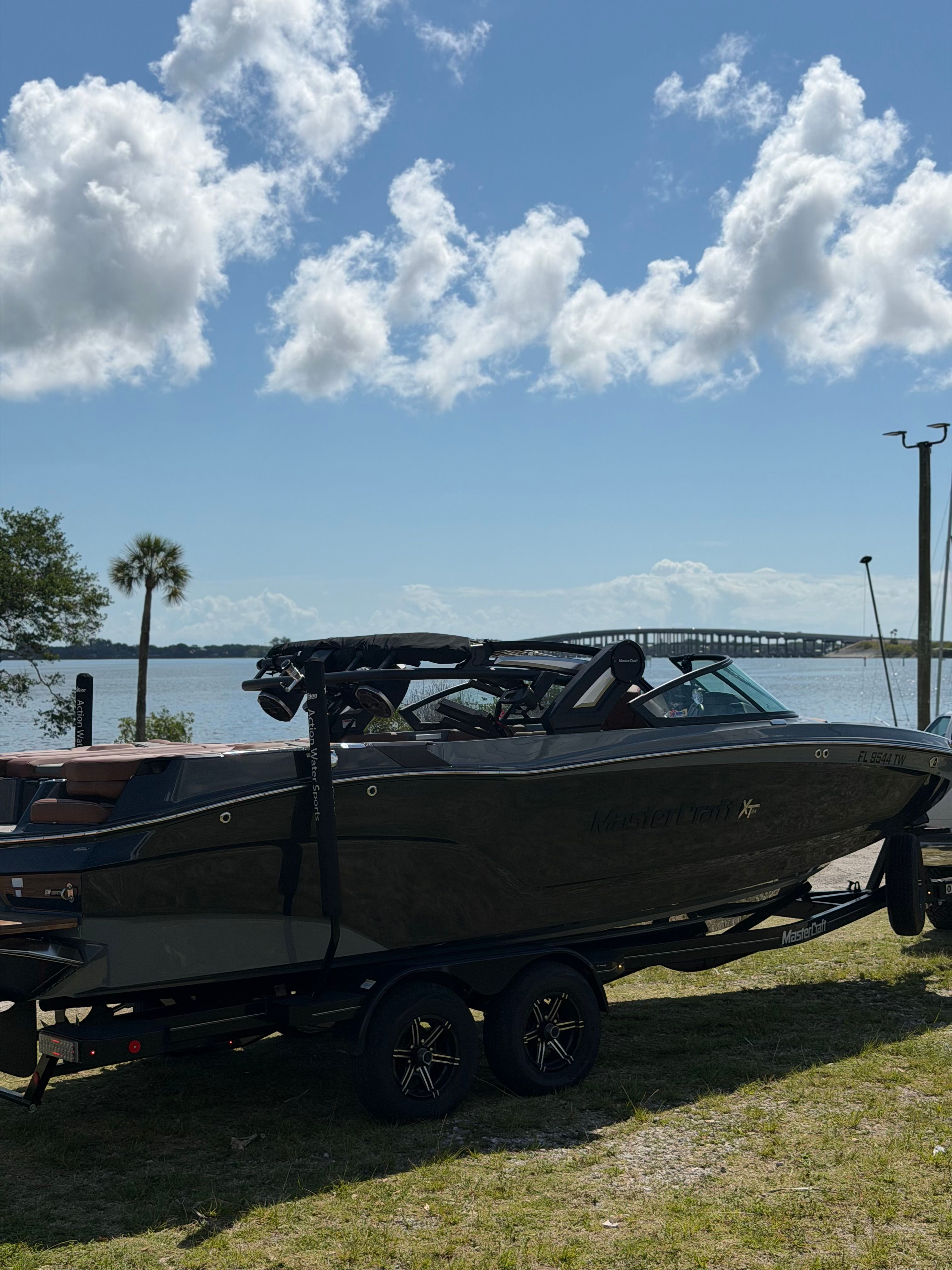 A MasterCraft XT24 boat on a trailer by the shore with a bridge in the background.
