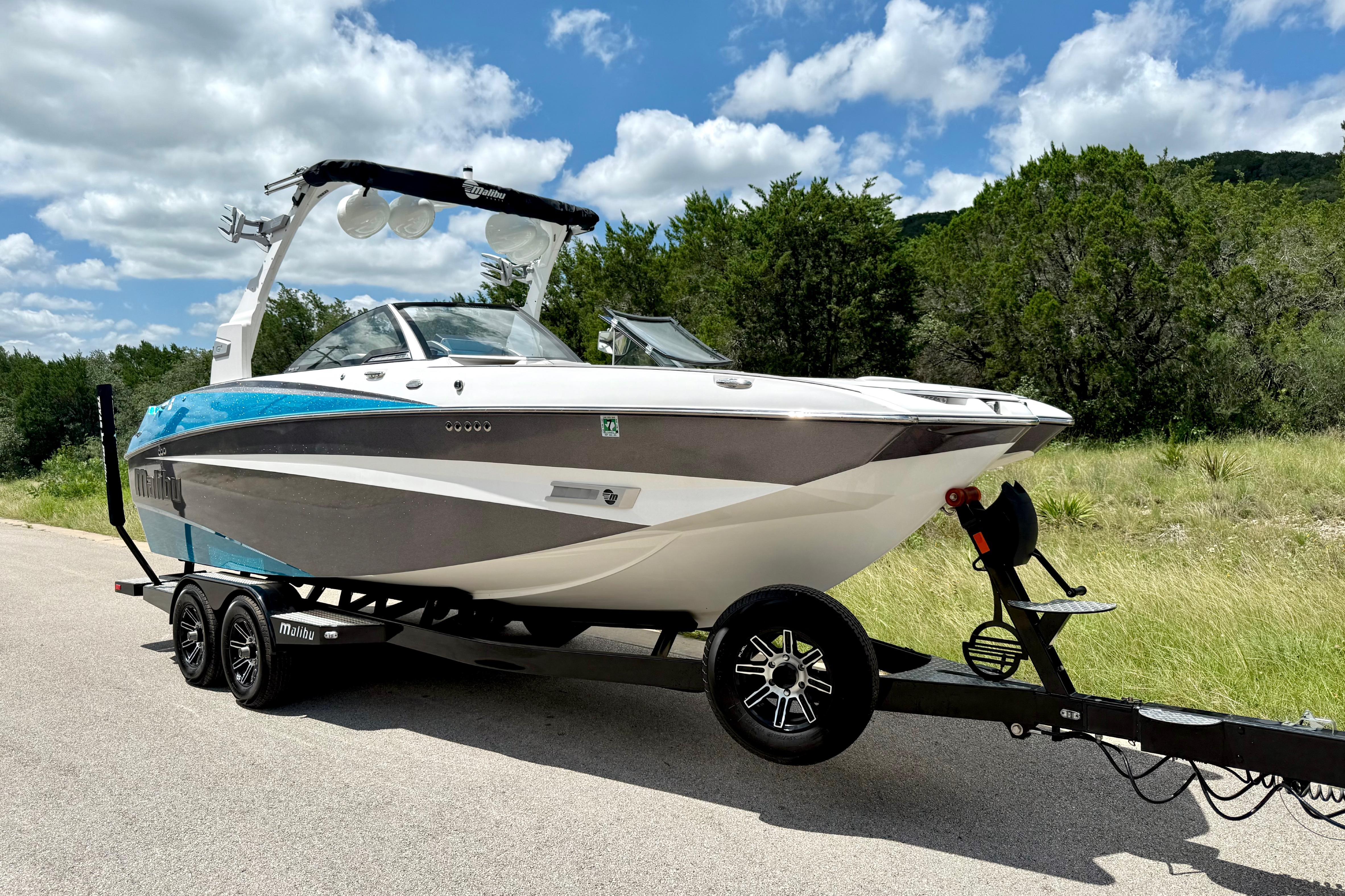 2017 Malibu M235 boat on trailer, parked on road with lush greenery and blue sky.