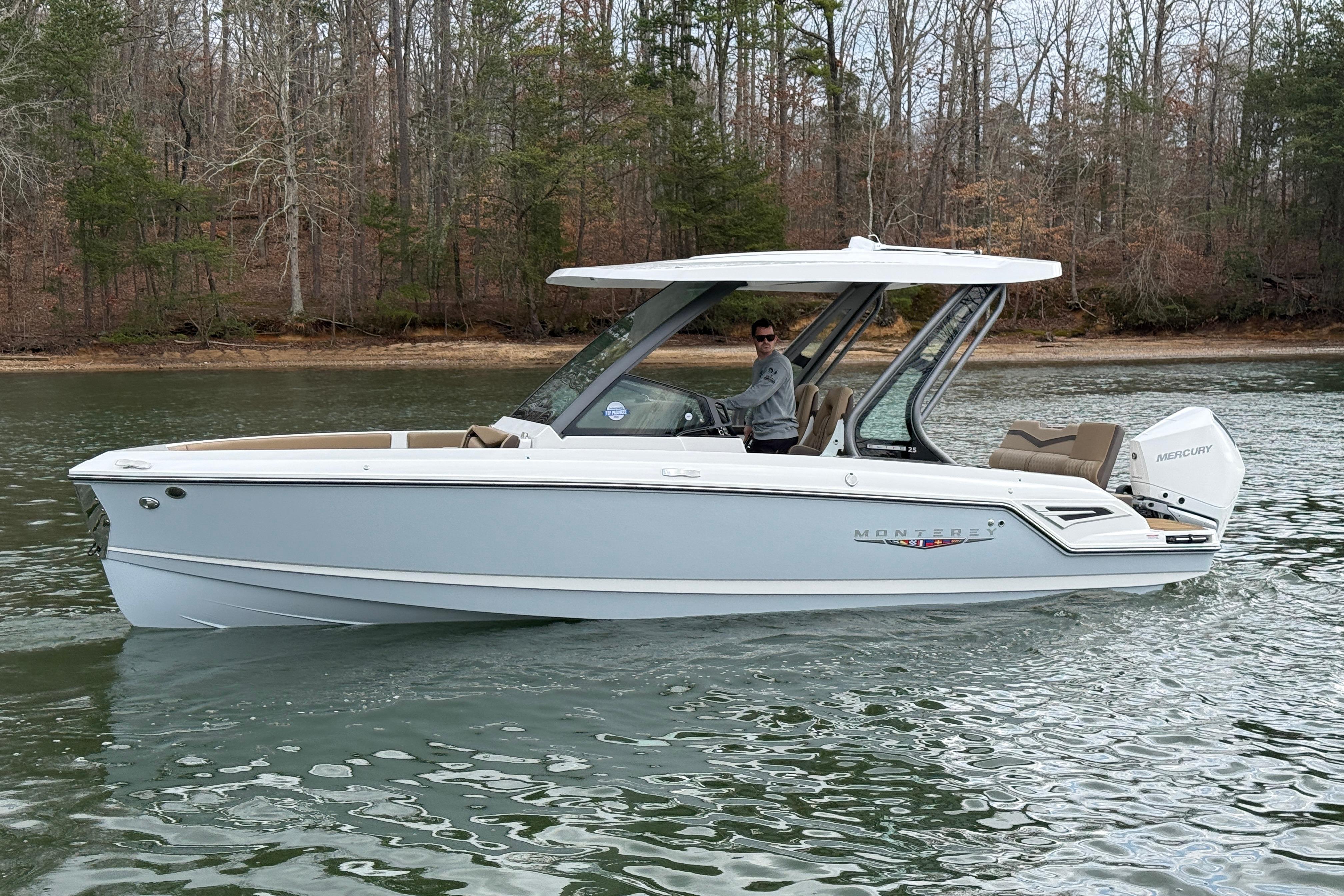 2026 Monterey Elite 25 OB boat cruising on a calm lake with wooded shoreline.
