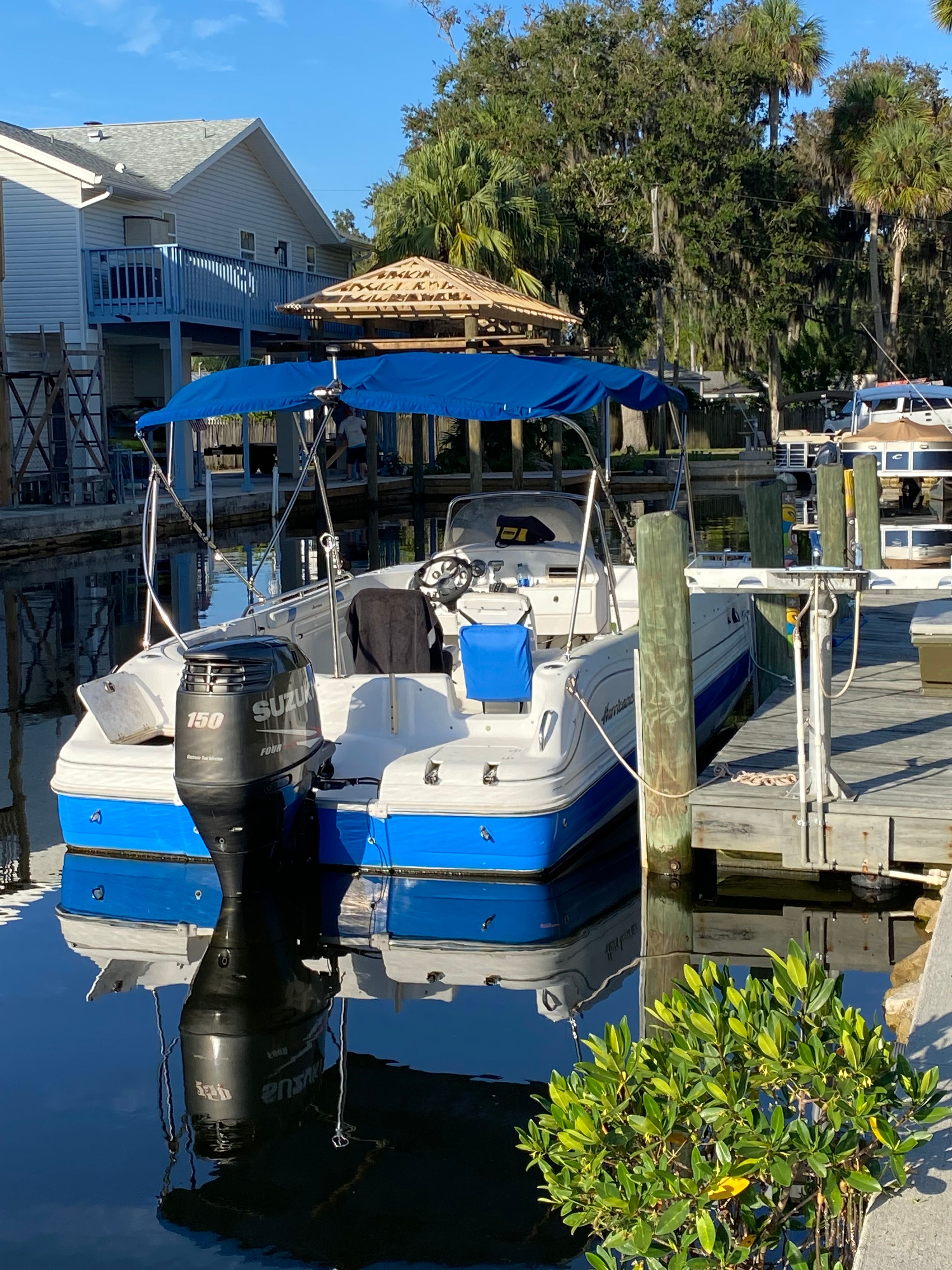 A blue and white boat with a sunshade docked in calm waters, surrounded by trees.