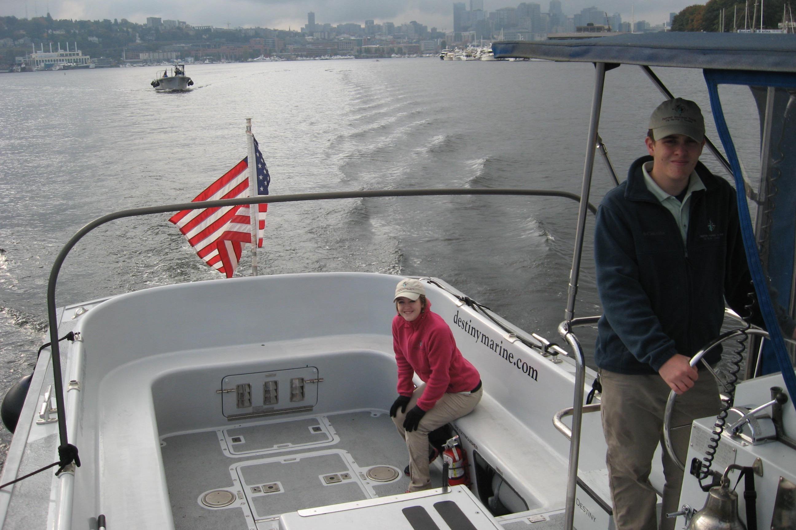 Two people on a 1994 Willard US Navy boat with American flag, city skyline in background.