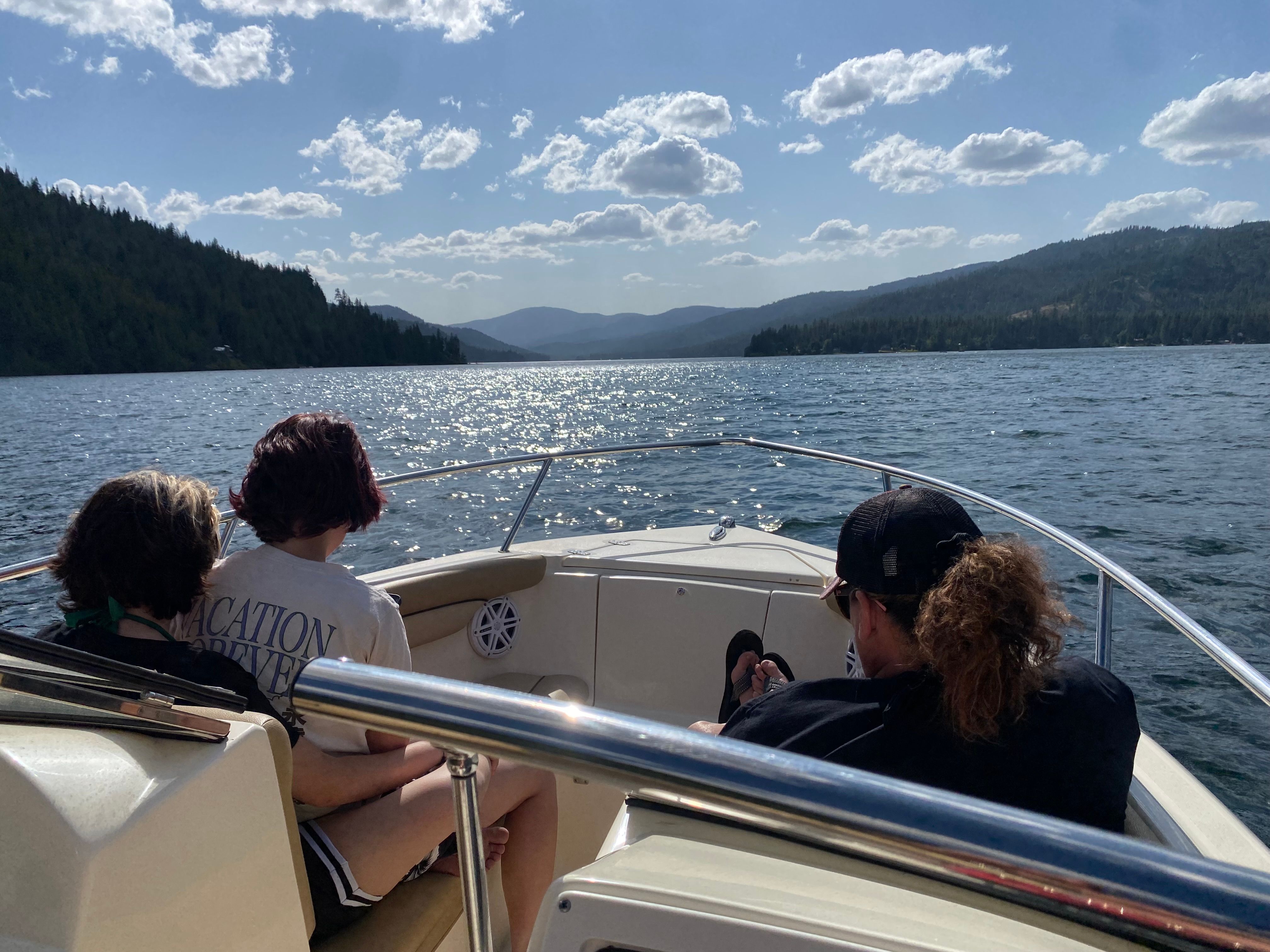 Two people sit on a boat cruising a calm lake, surrounded by wooded hills.
