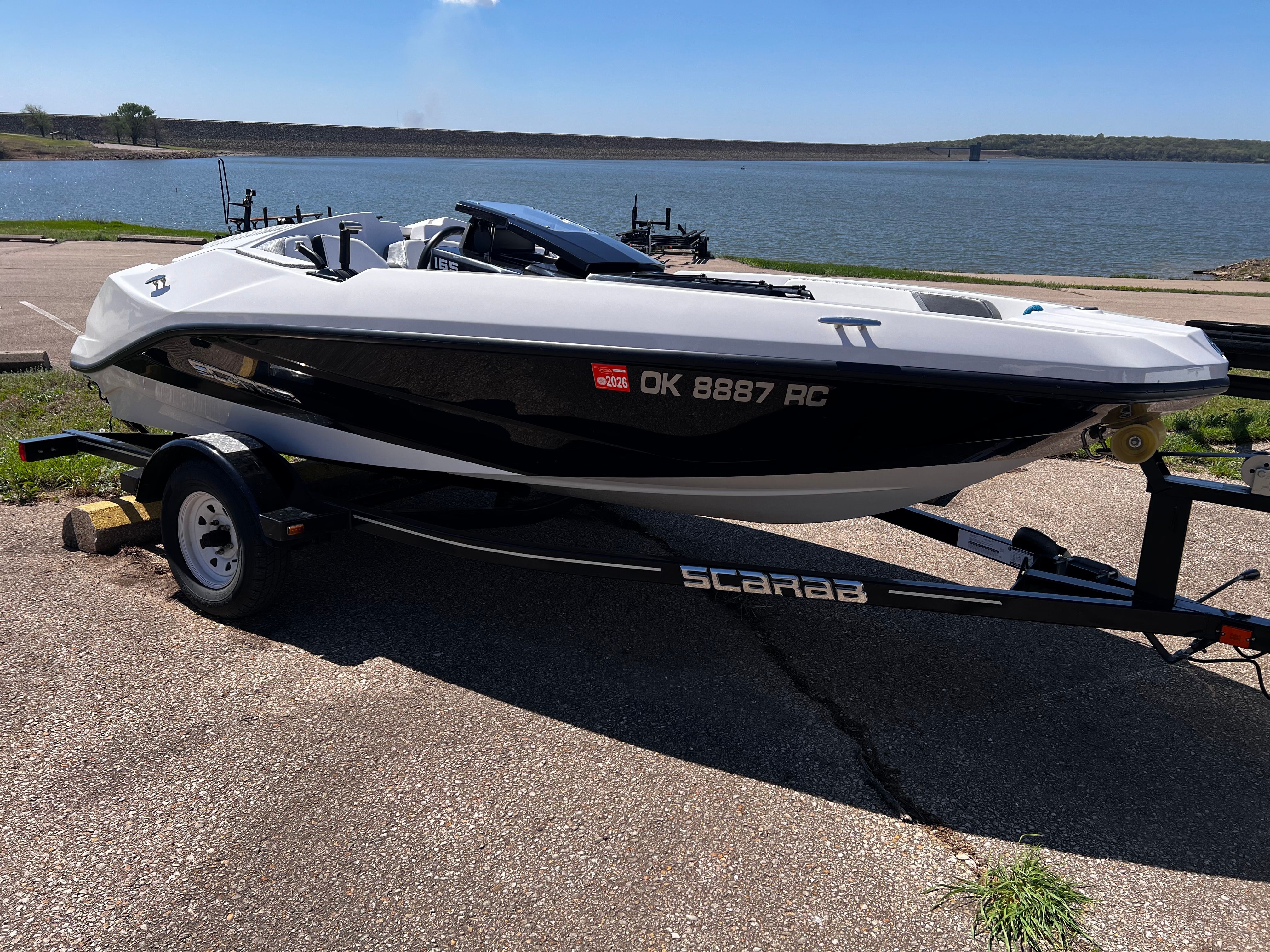 A white Scarab boat on a trailer, parked in a lot next to a lake.