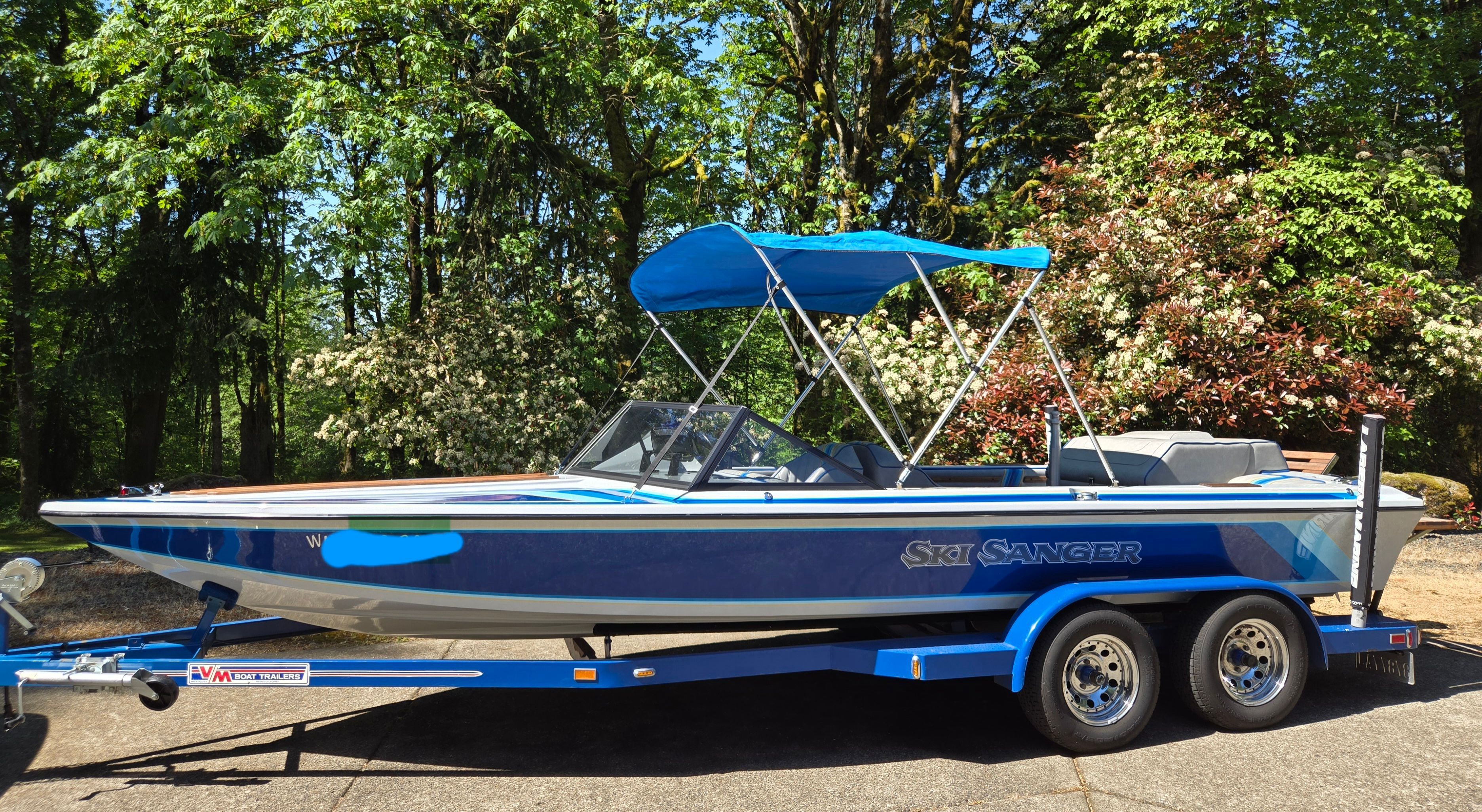A blue 1990 Sanger Ski Sanger on a trailer, surrounded by trees.