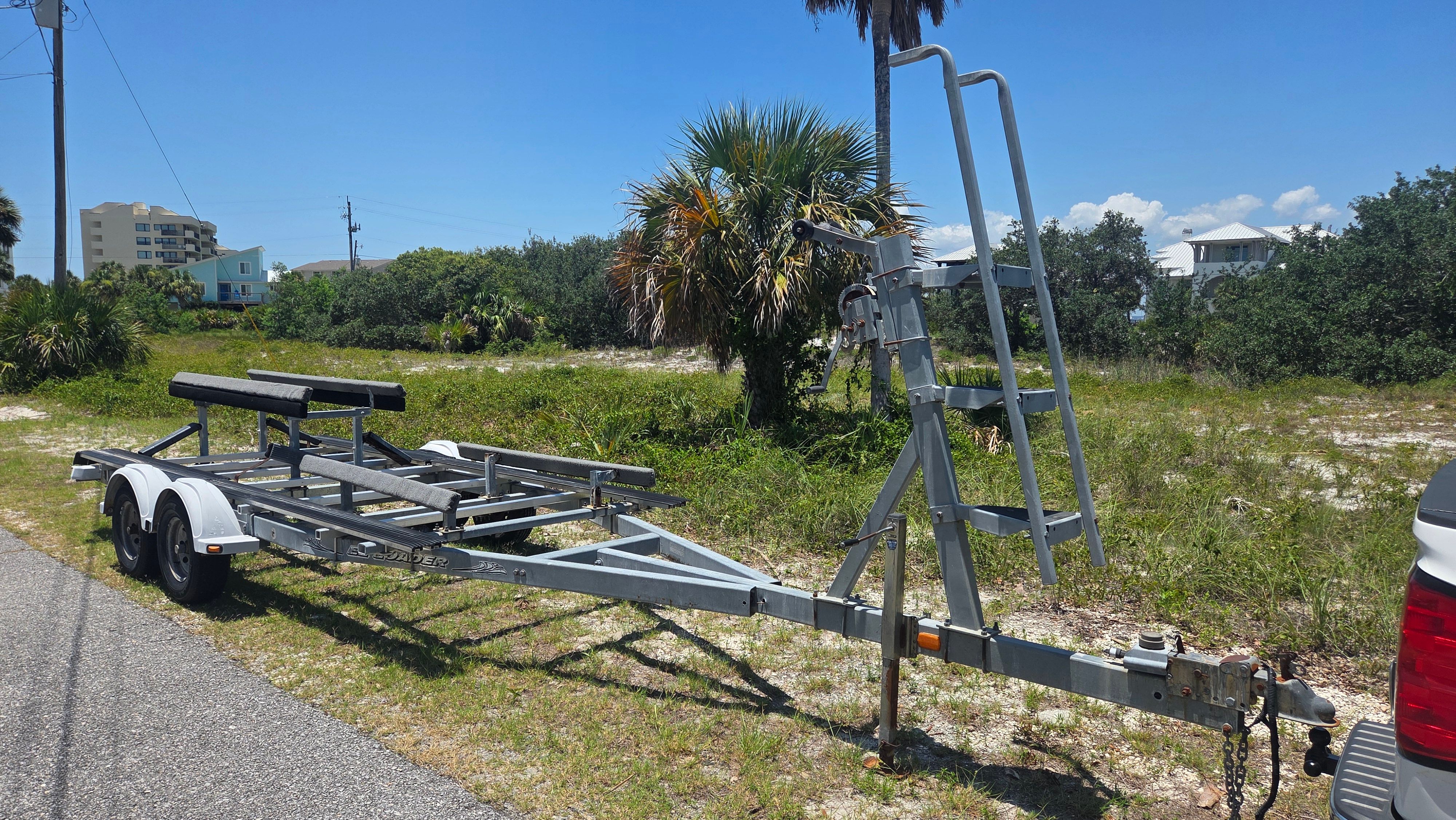 A boat trailer is parked on a road next to a grassy area with palm trees.