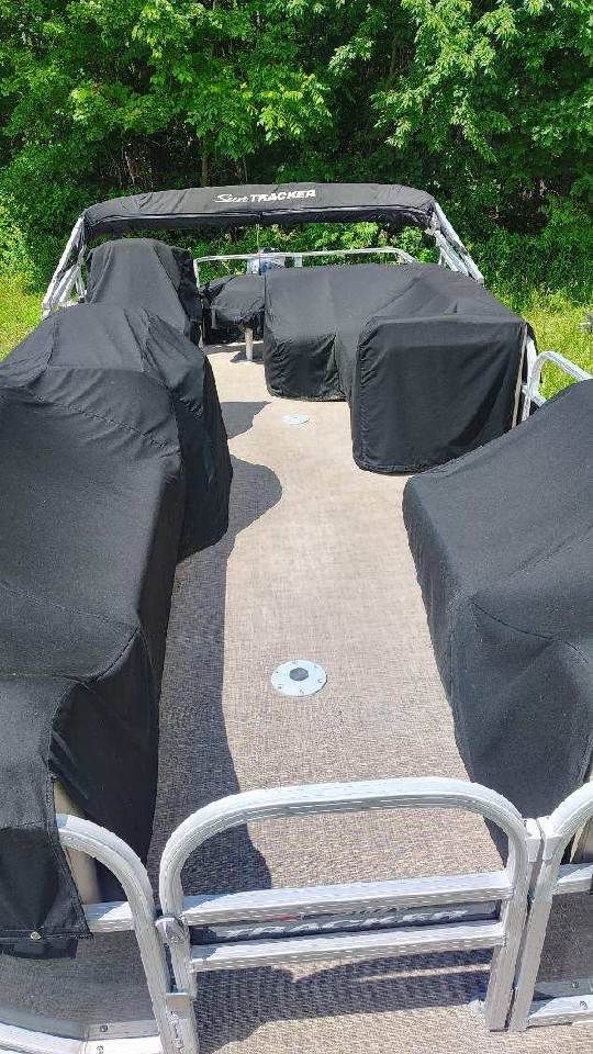 A pontoon boat with black covers on the seats, surrounded by green vegetation.