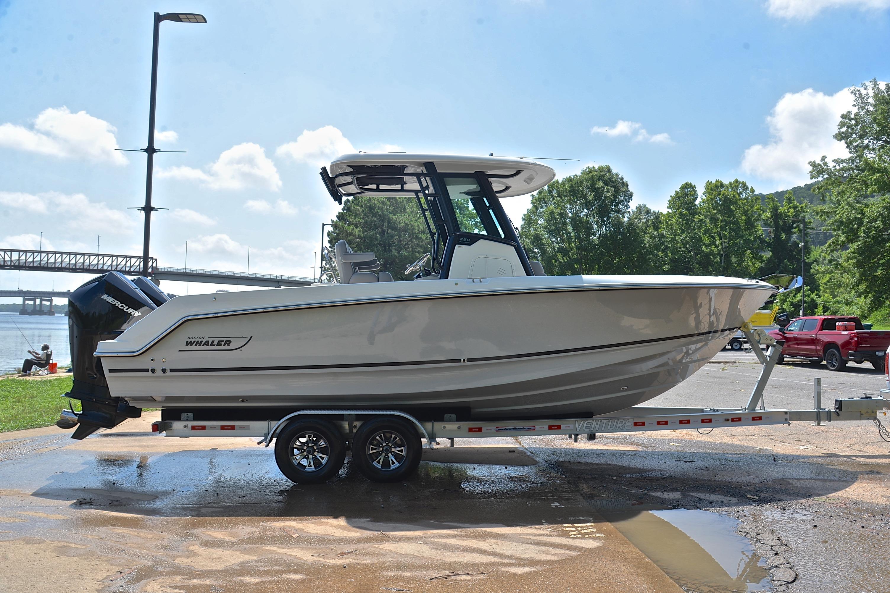 2024 Boston Whaler 250 Outrage boat on trailer, parked near a river with trees and bridge.