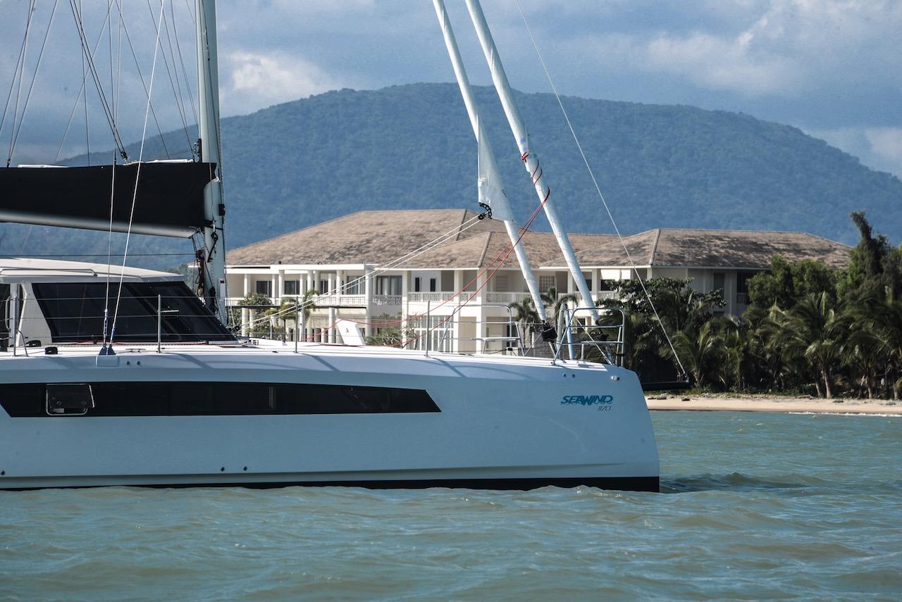 2025 Seawind 1170 catamaran sailing near tropical resort with mountain backdrop.