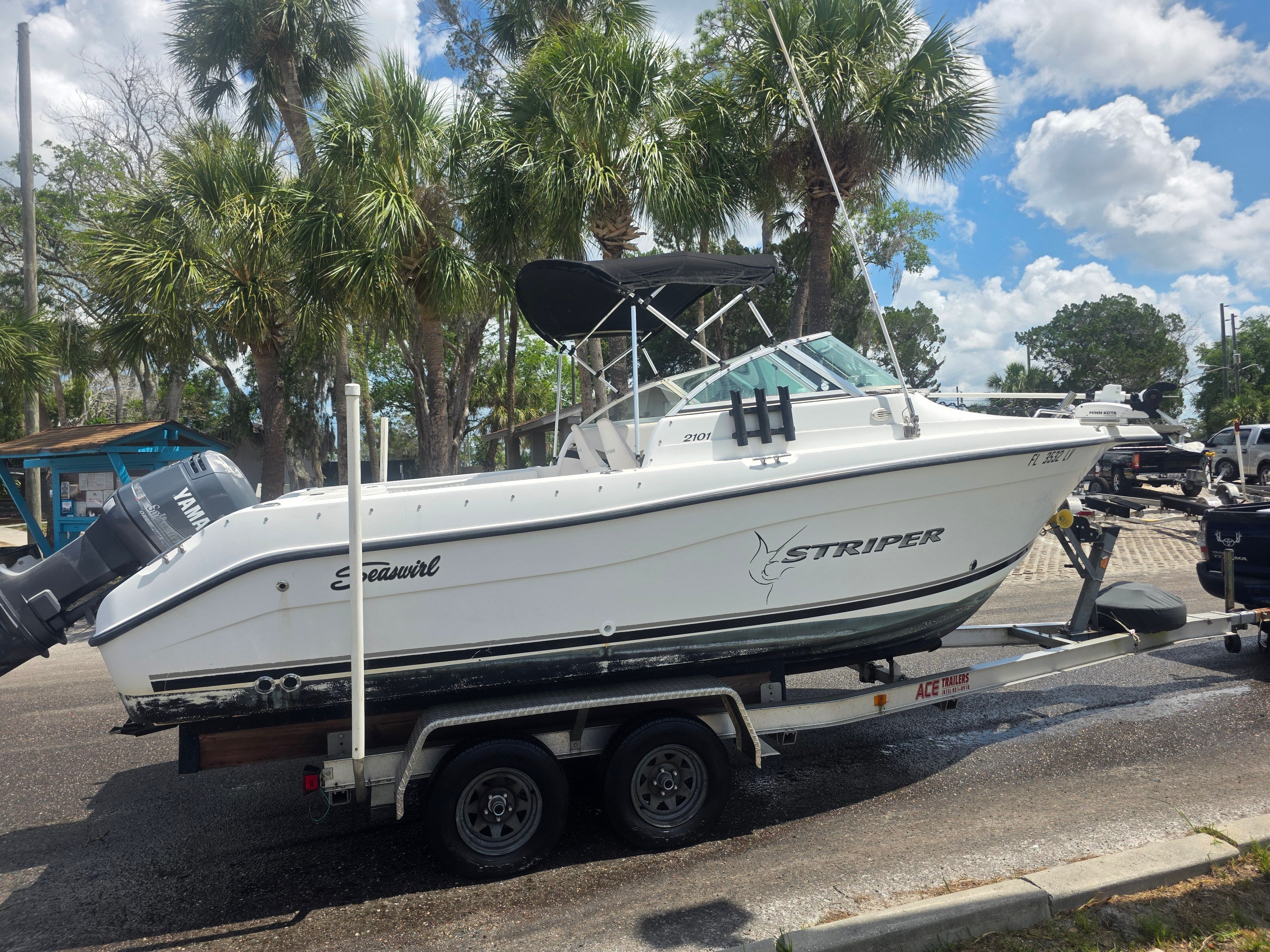A white boat on a trailer, surrounded by palm trees and a cloudy sky.