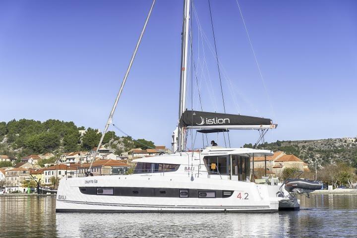 2023 Bali 4.2 catamaran sailing near a coastal village, clear blue sky.