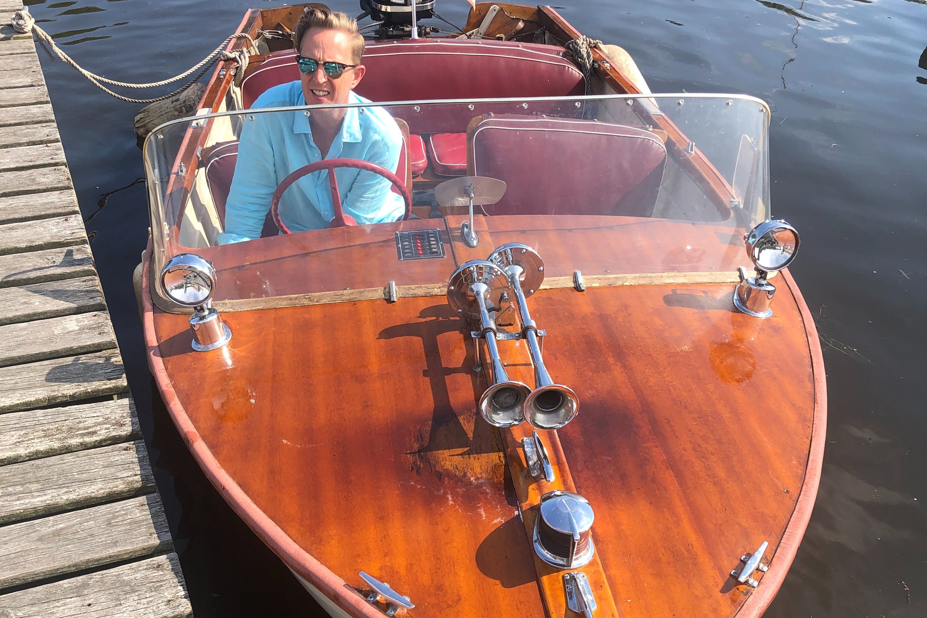 A classic wooden boat, MFG Cambridge from 1957, with red interior and a man at the helm.