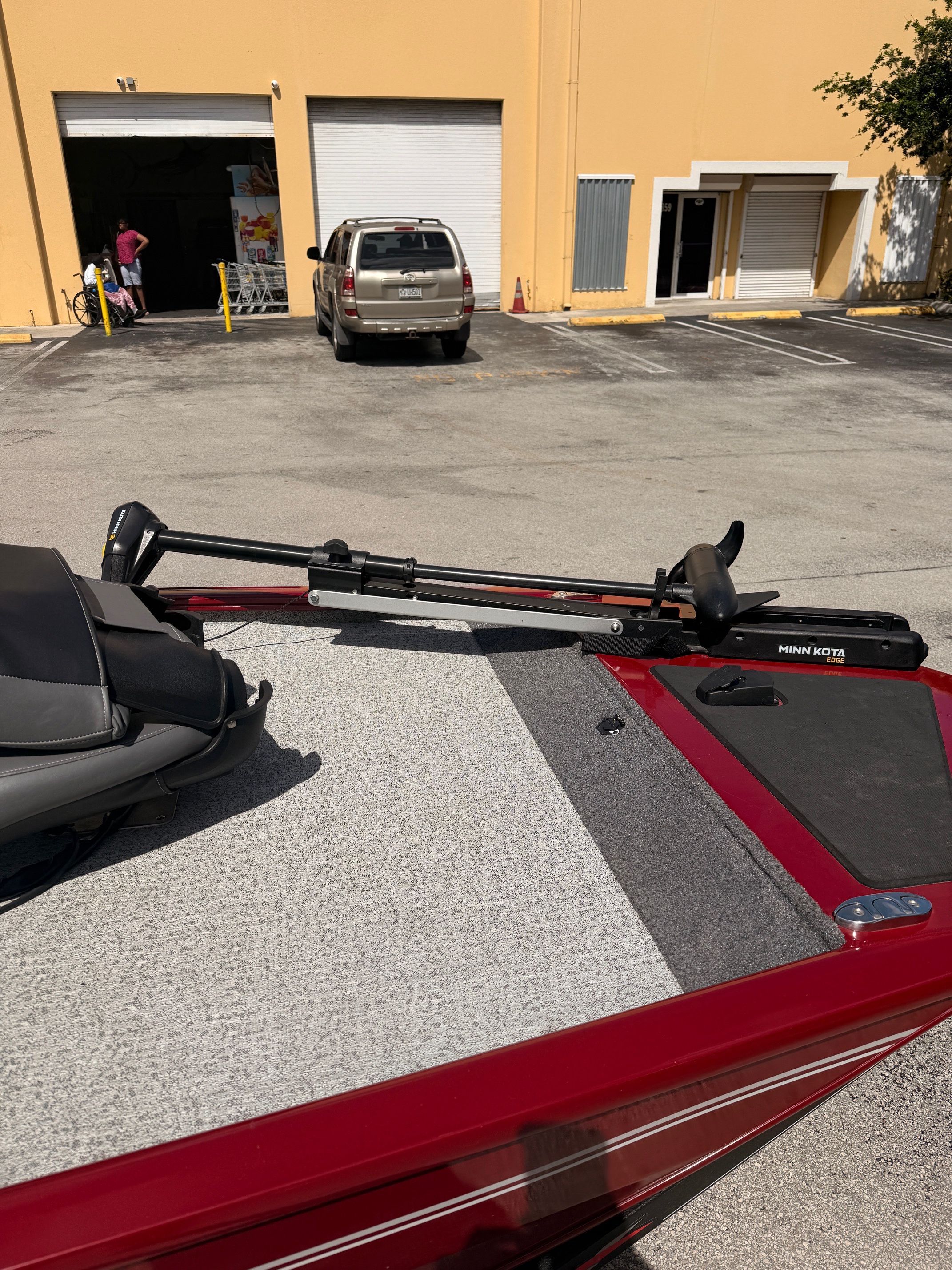 A red boat with gray carpeting is parked in a lot in front of a building.