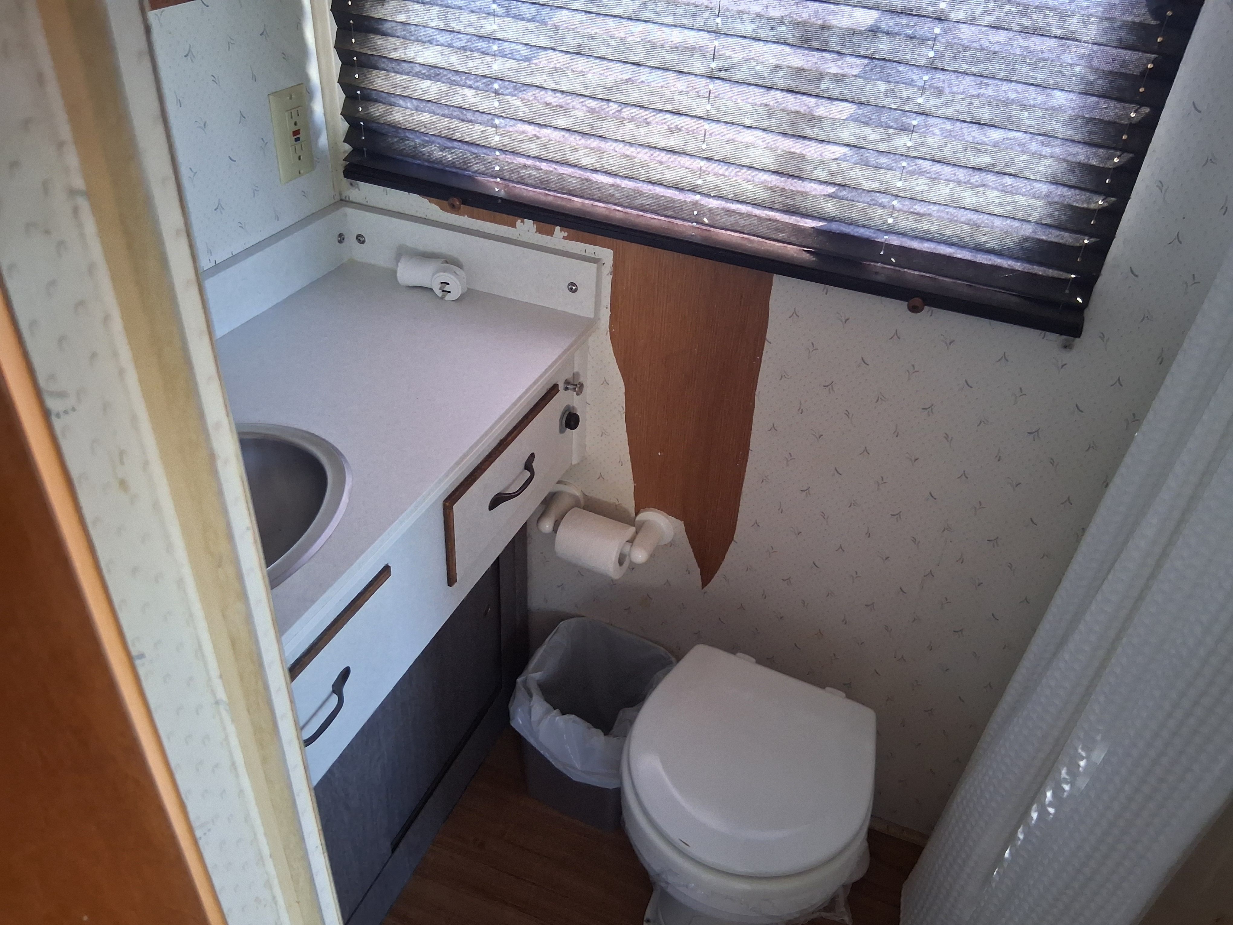 The image shows a bathroom with a toilet, sink, and a window with blinds.