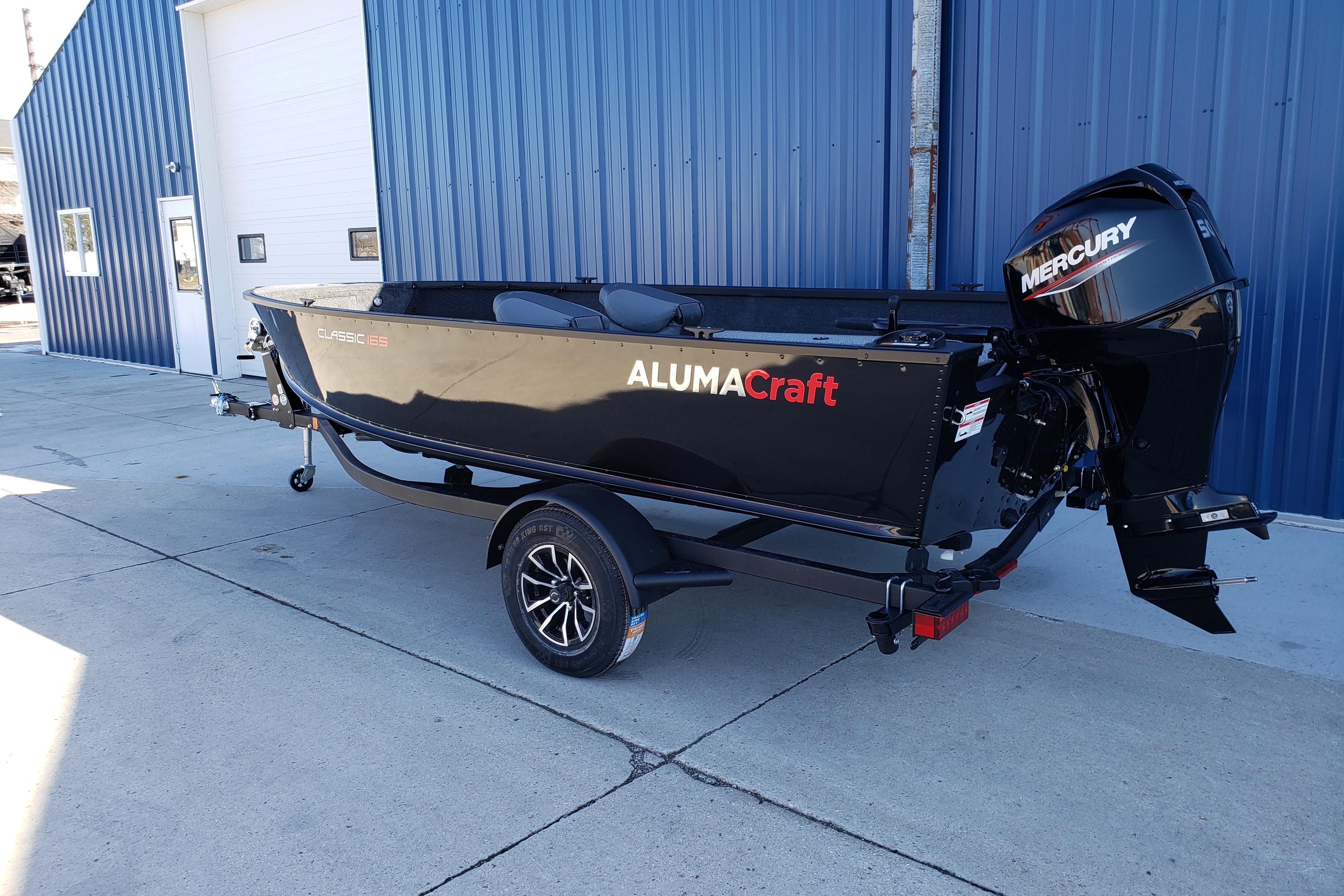 2026 Alumacraft Classic 165 Tiller boat with Mercury engine, parked outside a blue building.