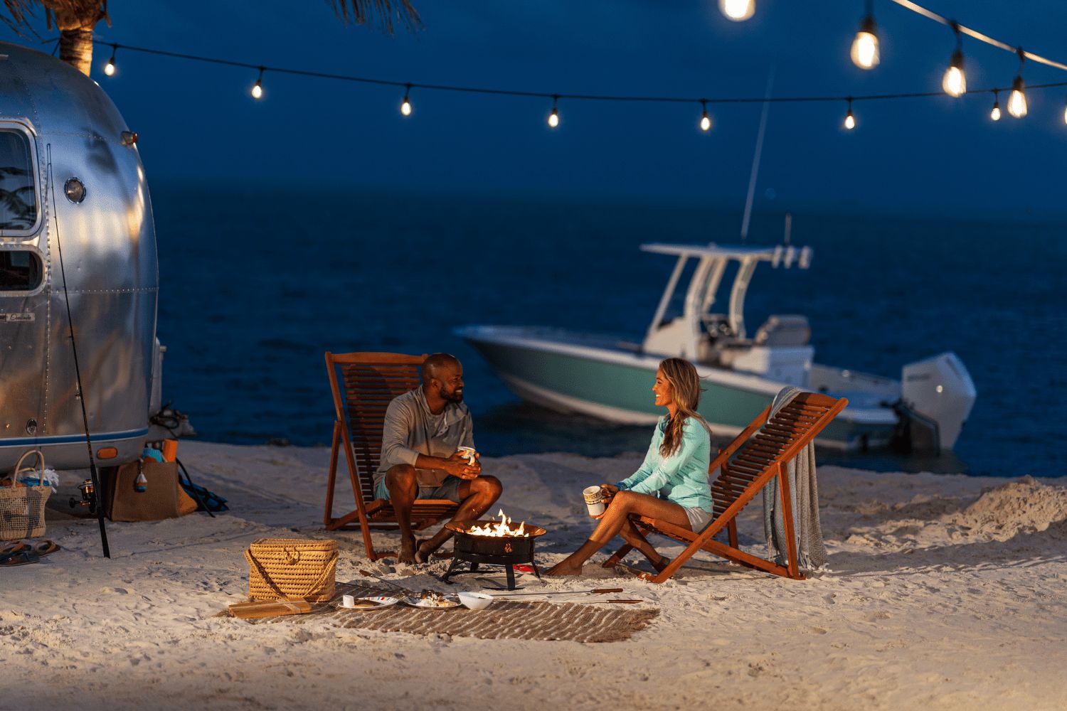 A couple sits by a beach campfire, with a Boston Whaler 25DA in the background.