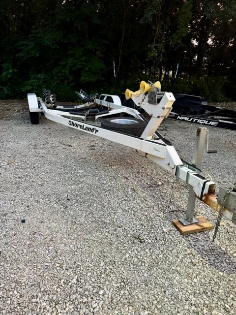 A boat trailer sits on a gravel lot, surrounded by trees and other trailers.