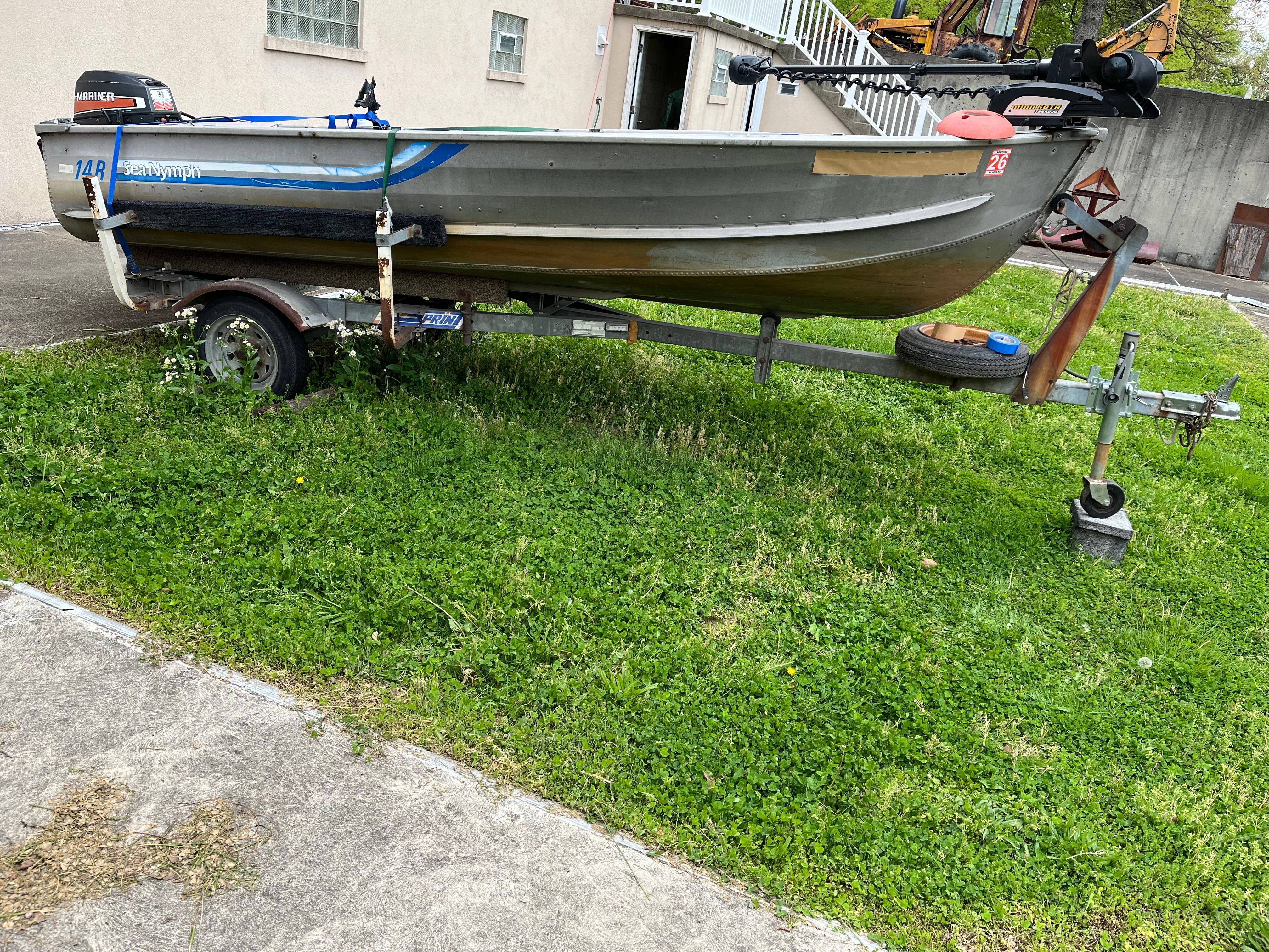 A gray boat on a trailer sits on grass beside a building.