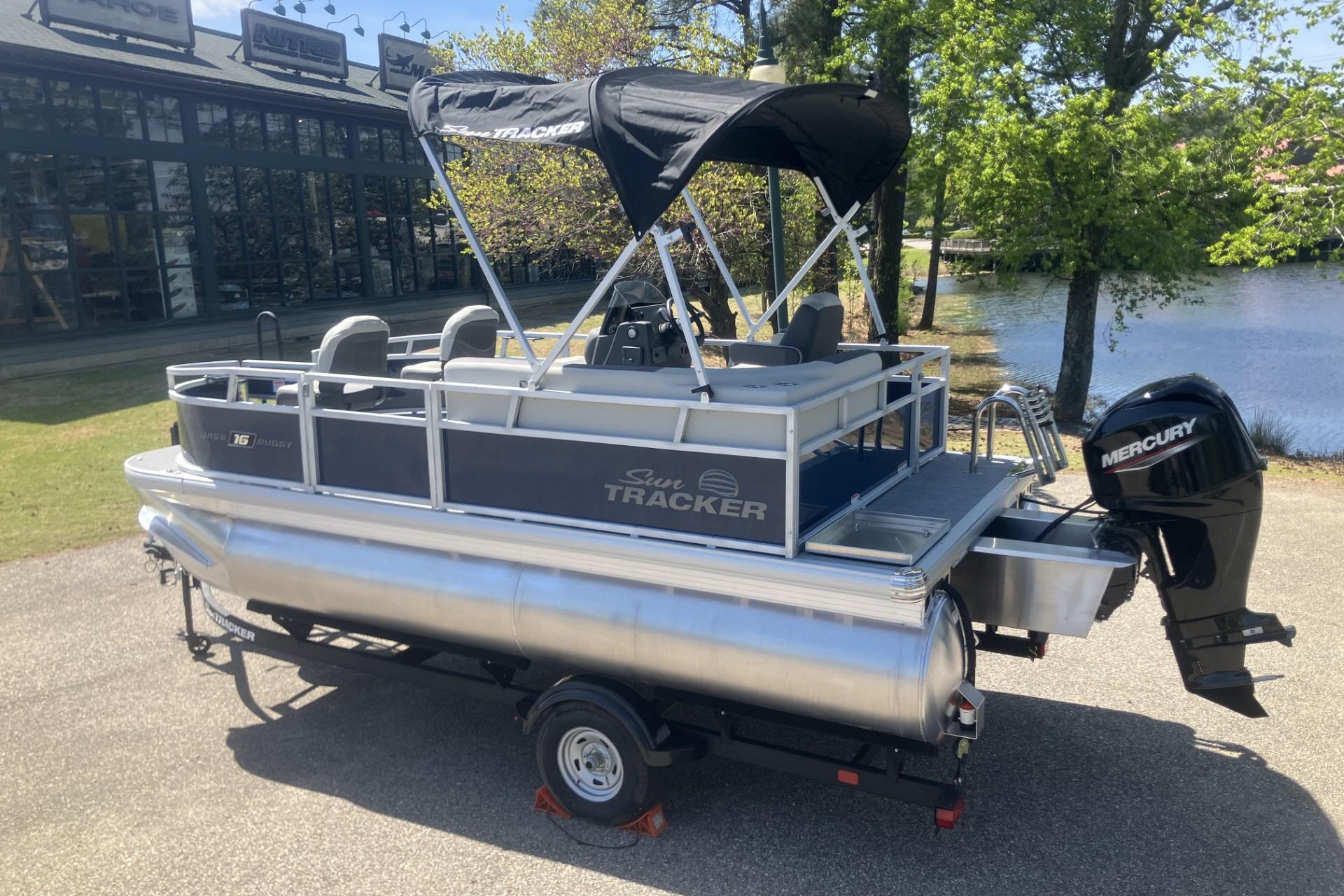 2026 Sun Tracker Bass Buggy 16 XL Select pontoon boat with Mercury engine, parked near water.