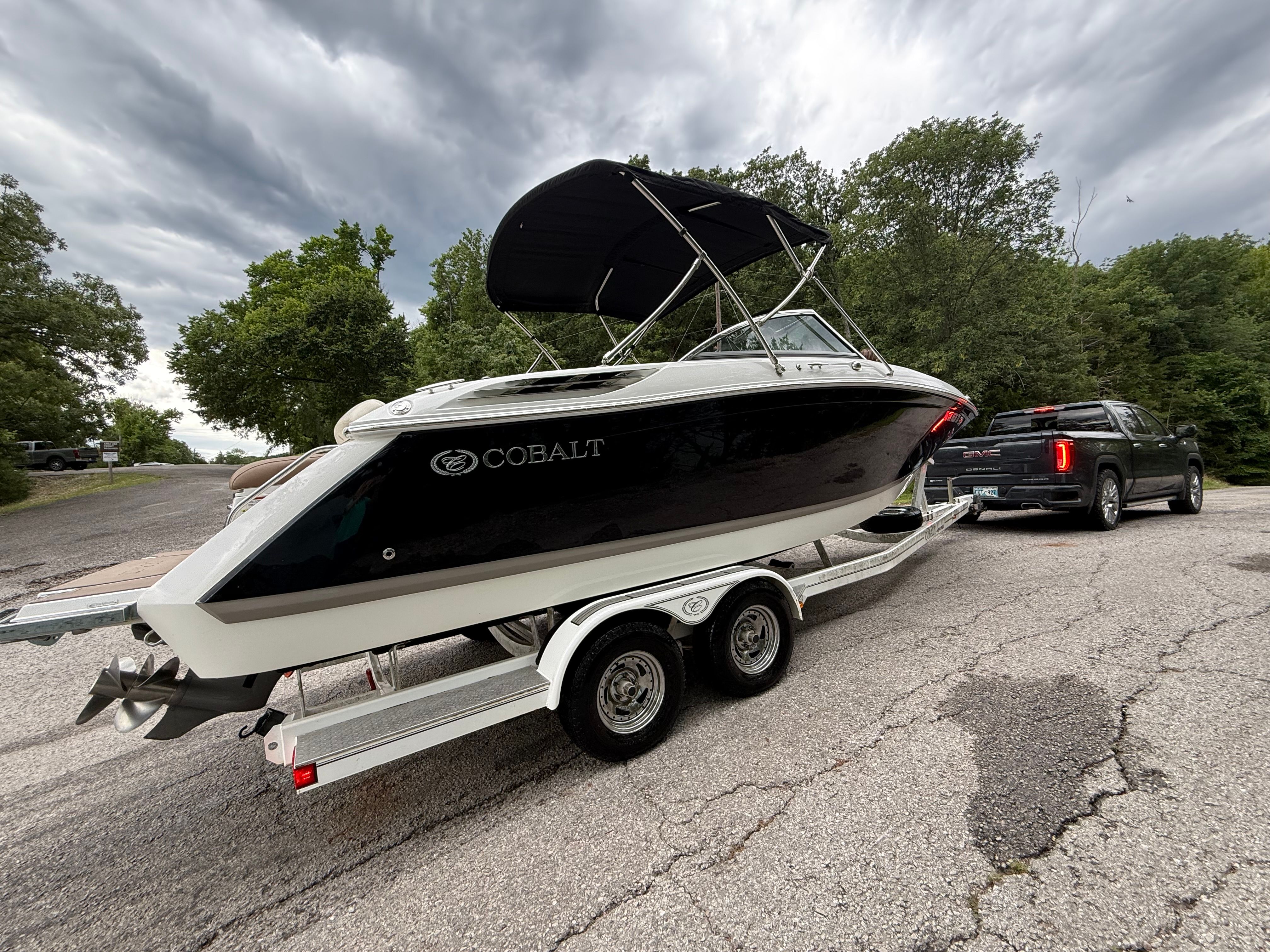 A Cobalt boat on a trailer, towed by a black vehicle, surrounded by trees.