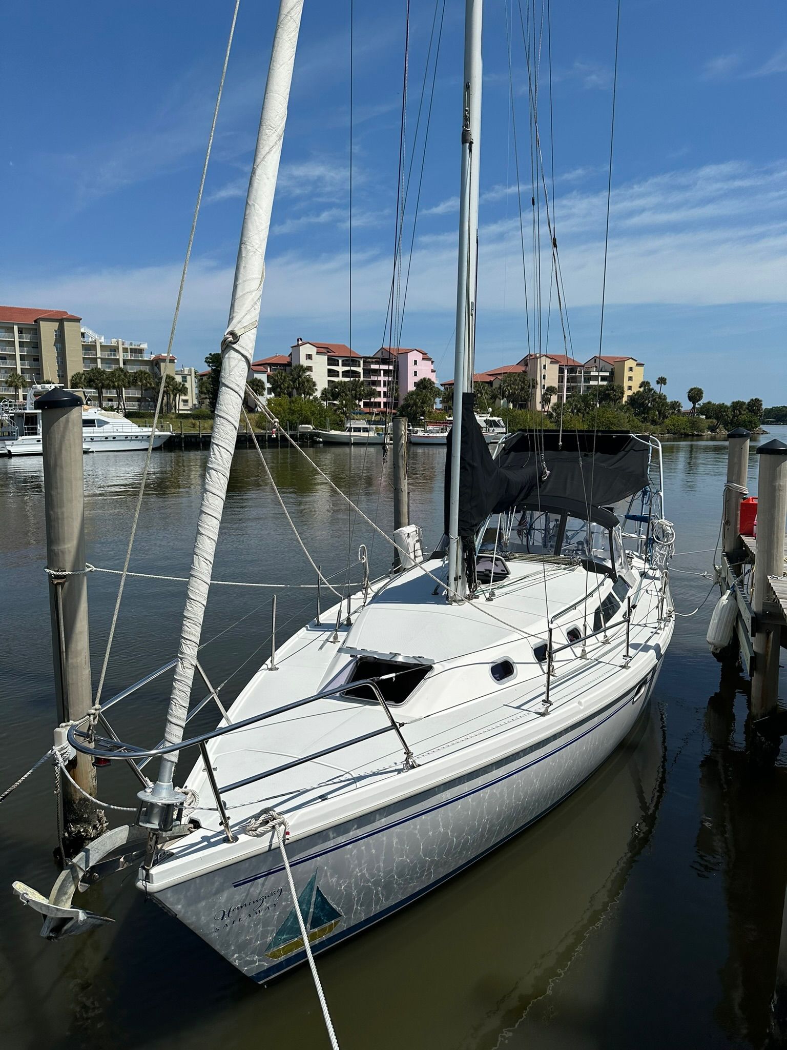 A white sailboat is docked in a calm harbor, surrounded by buildings and clear skies.