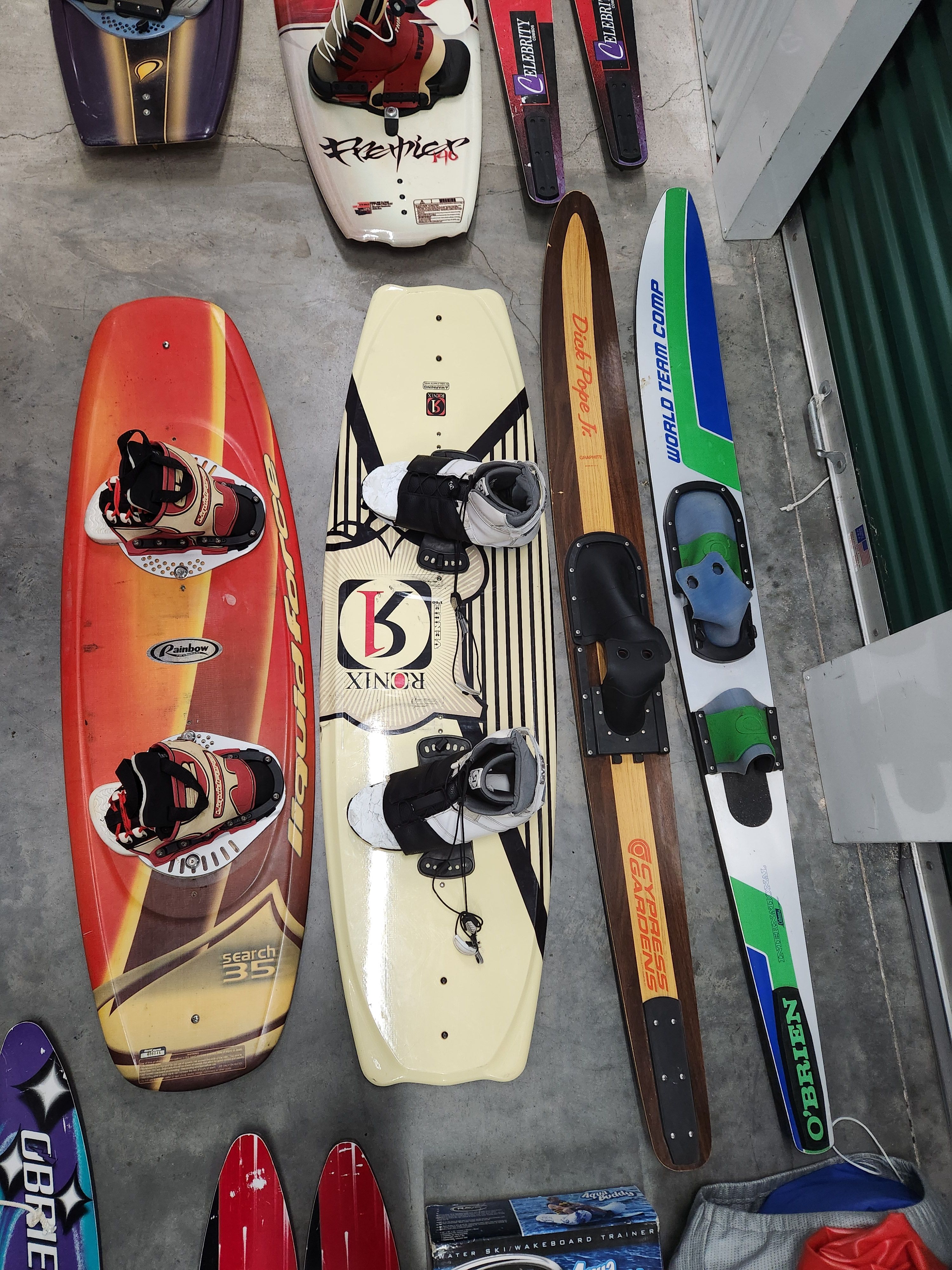 Various wakeboards and water skis on concrete floor.