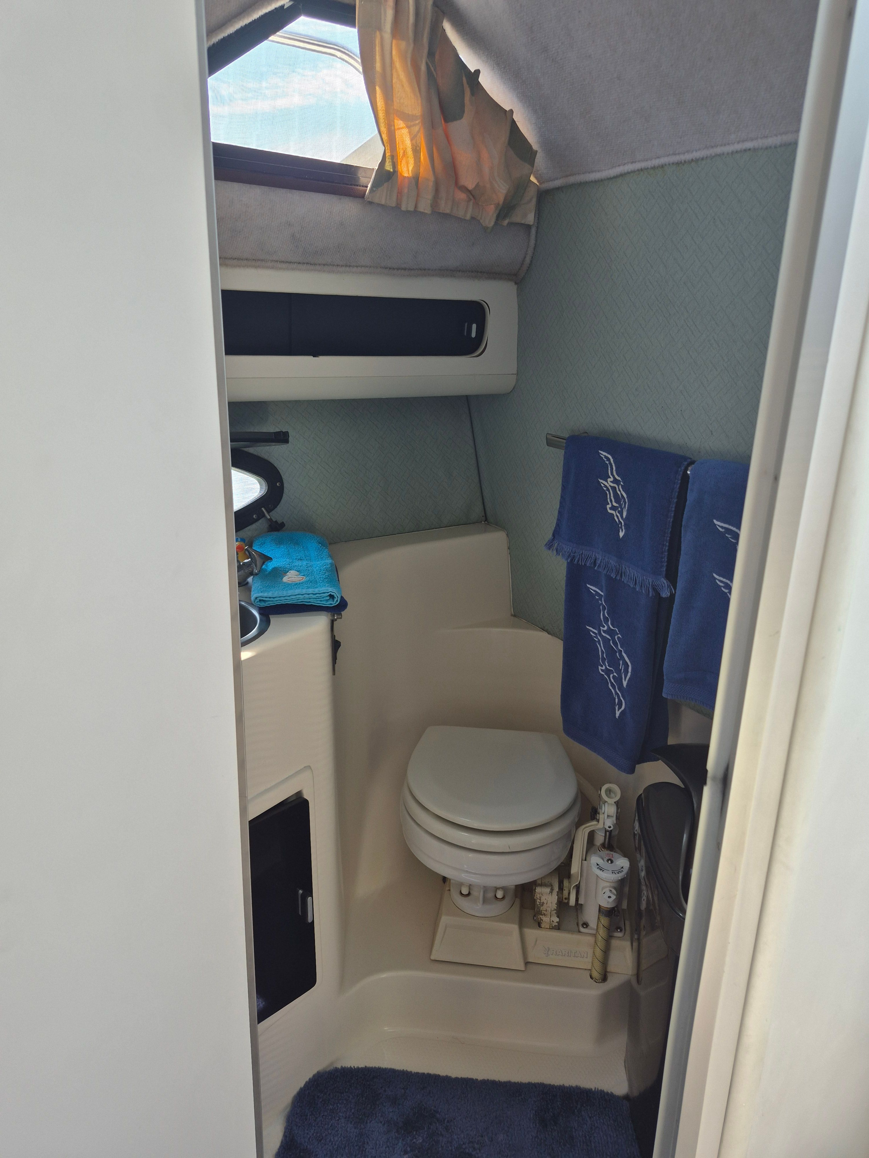 Interior view of a boat with toilet, sink, and blue towels. Window above.
