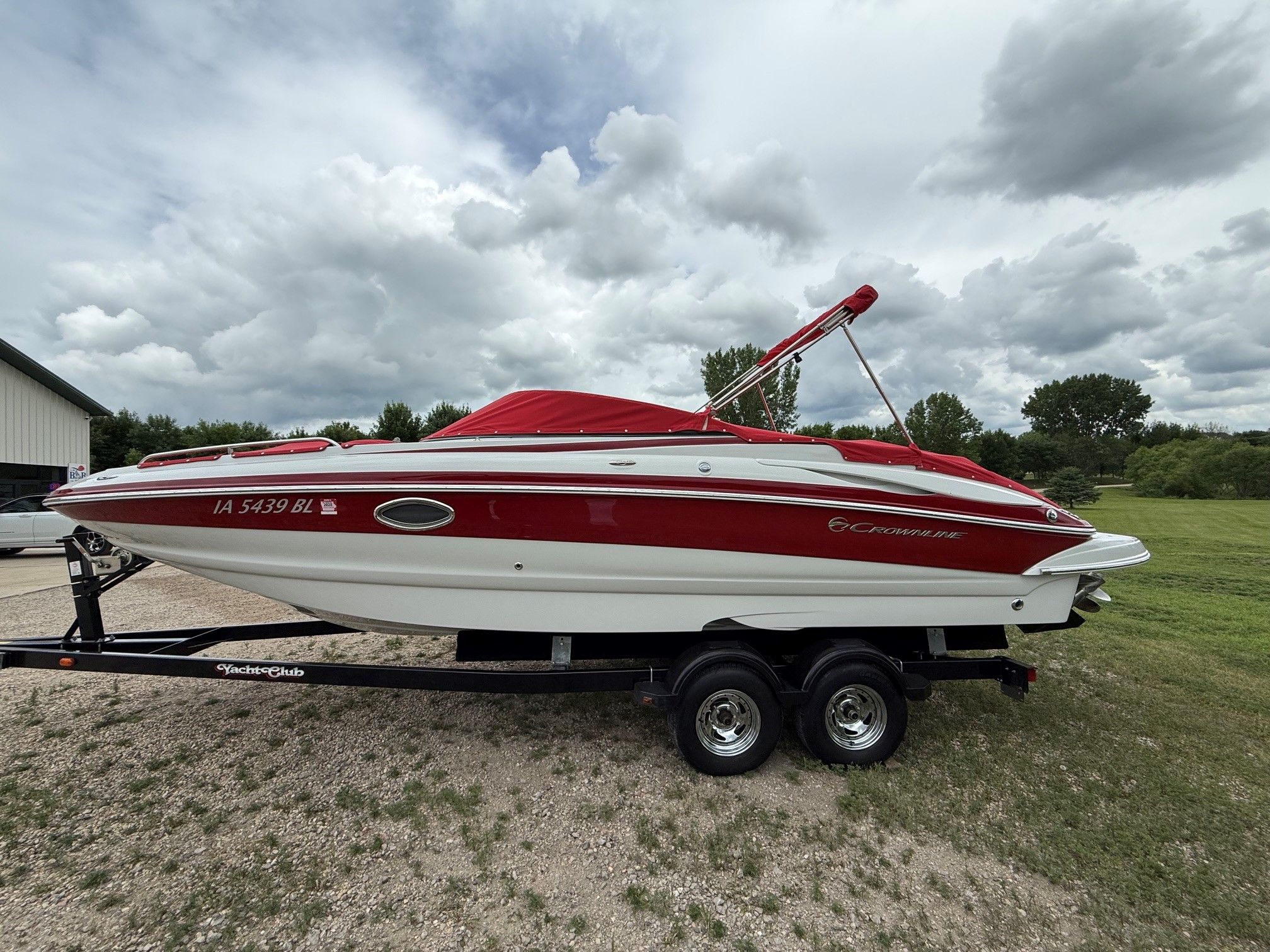 A 2012 Crownline 252 EX boat on a trailer, with a red cover, on grassy terrain.