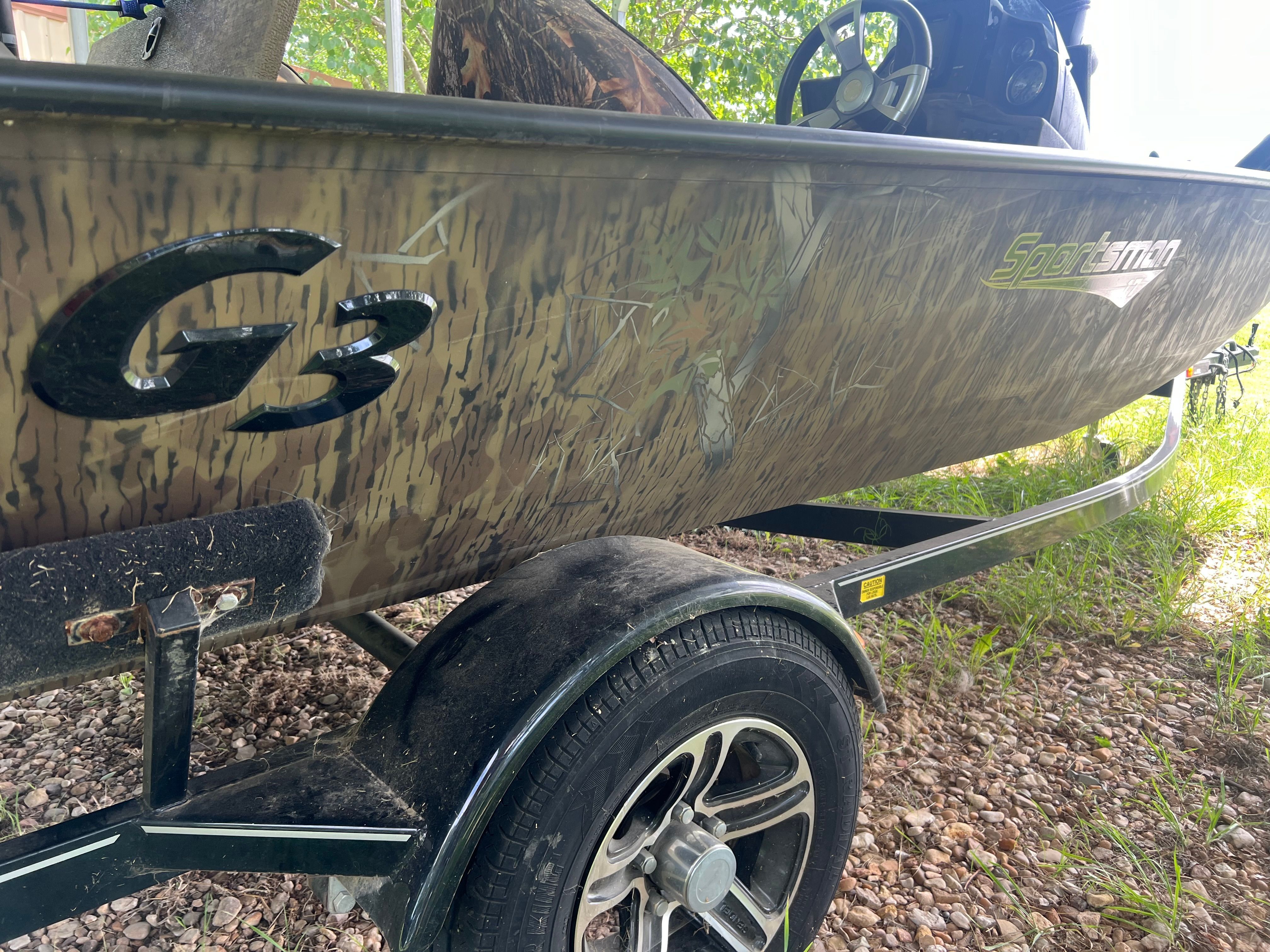 A 2017 G3 Sportsman 17 Camo boat on a trailer, surrounded by grass and trees.