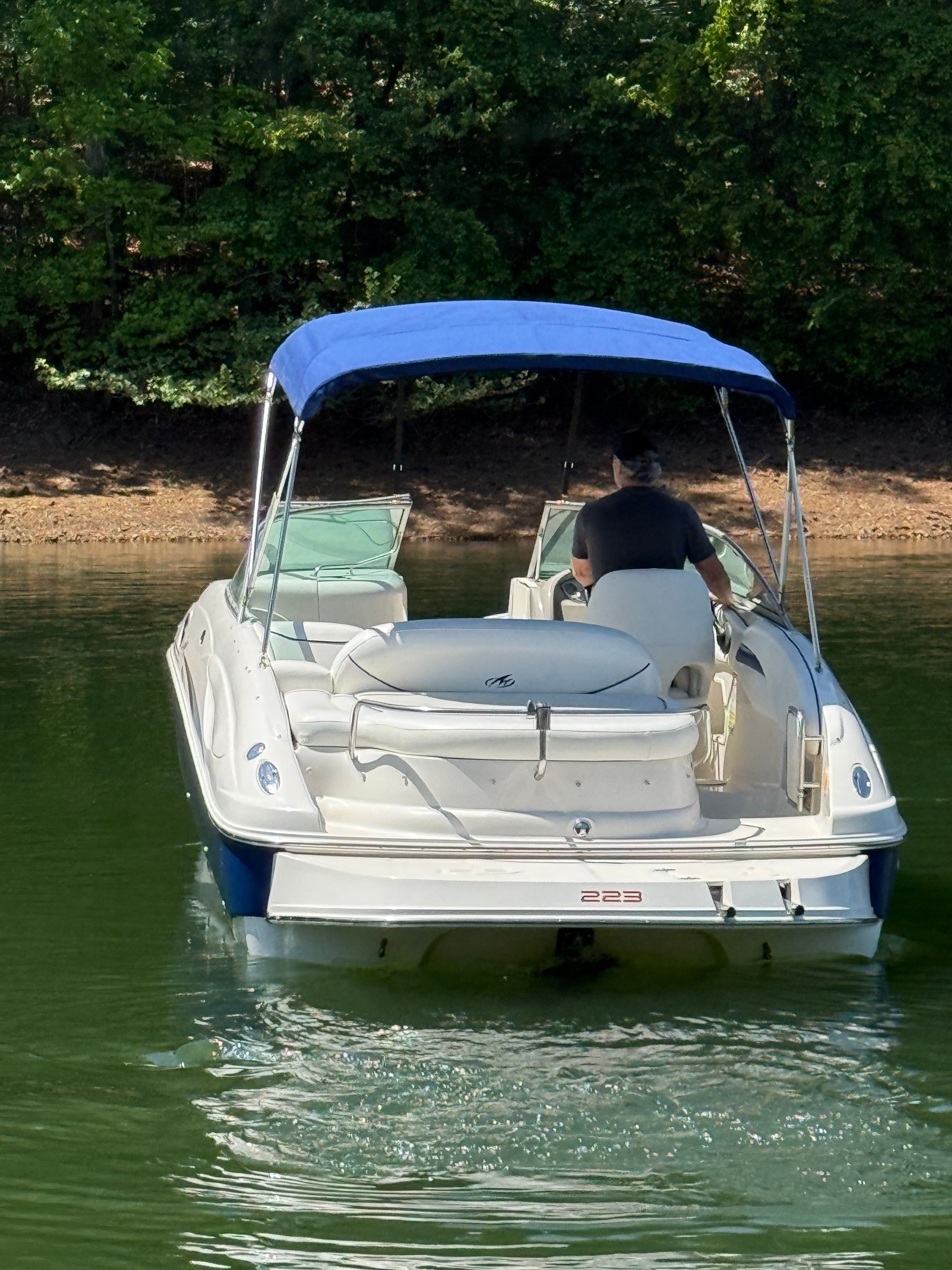 A white boat with a blue canopy navigates a calm lake, surrounded by trees.