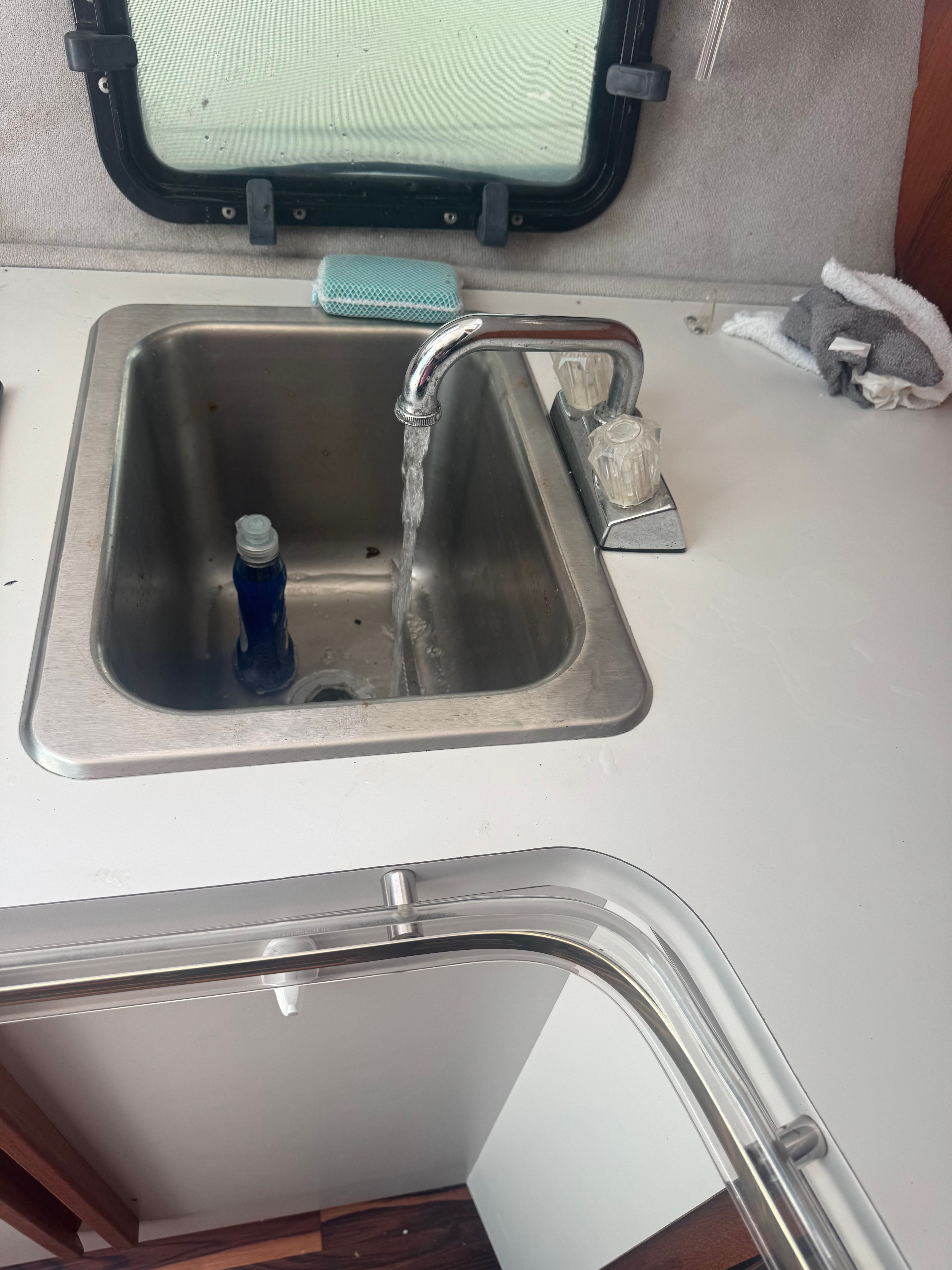 A sink with running water in a yacht galley, with a window and cleaning items nearby.