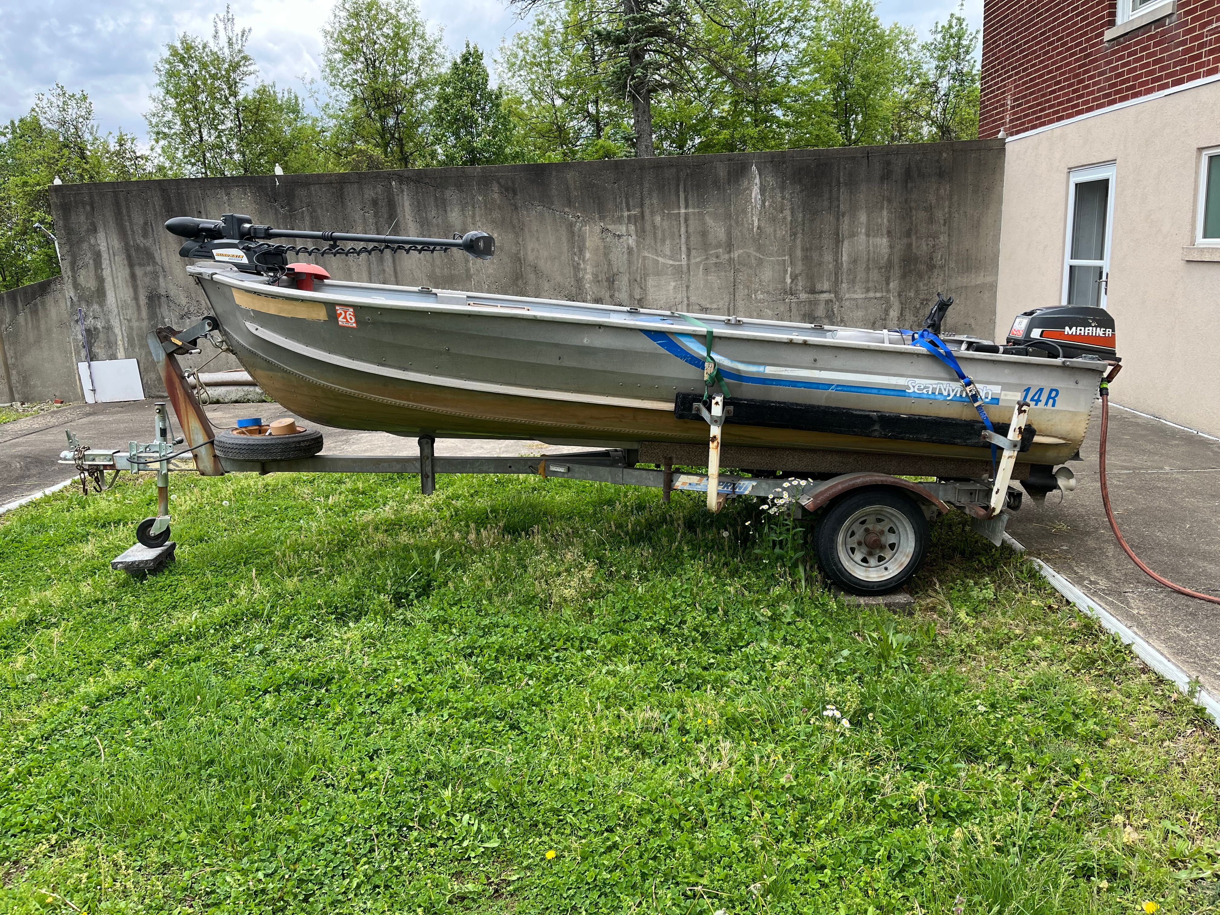 A 14-foot Sea Nymph boat on a trailer, parked on grass beside a building.